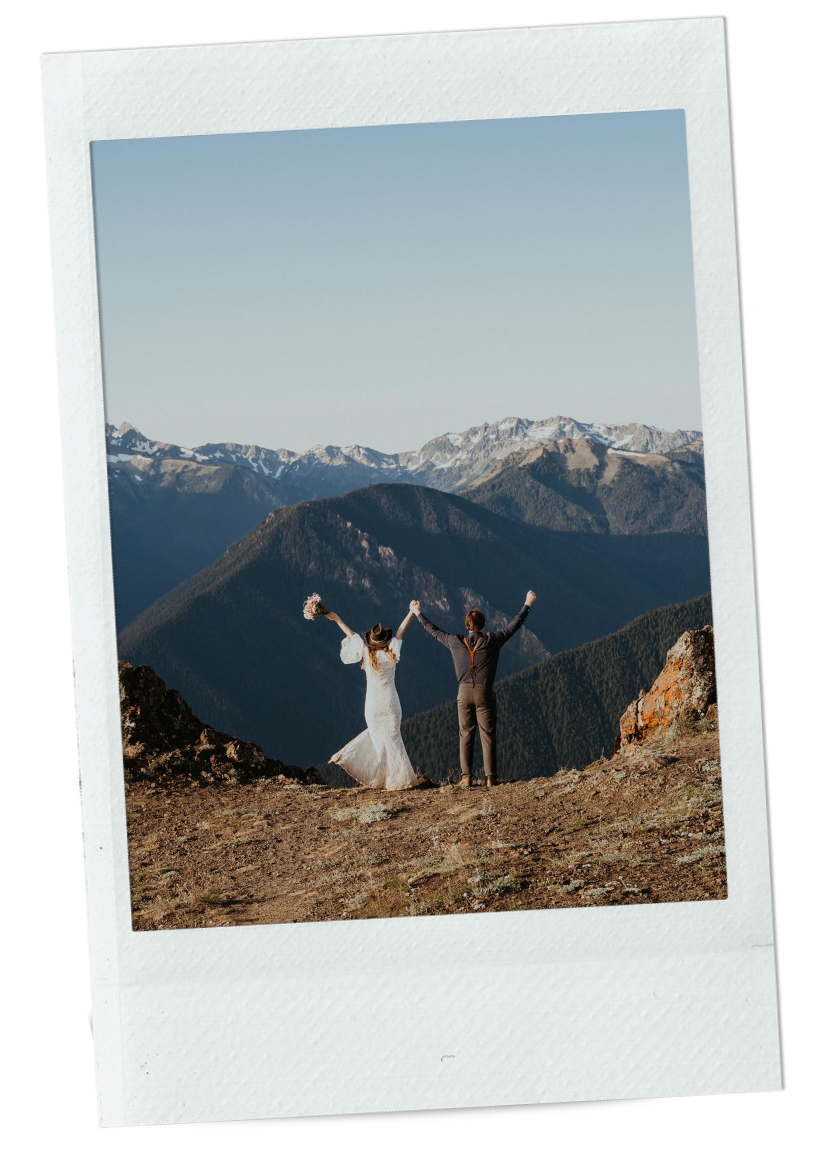 A bride and groom celebrating on a mountain overlook with a panoramic view of snow-capped peaks in the background.