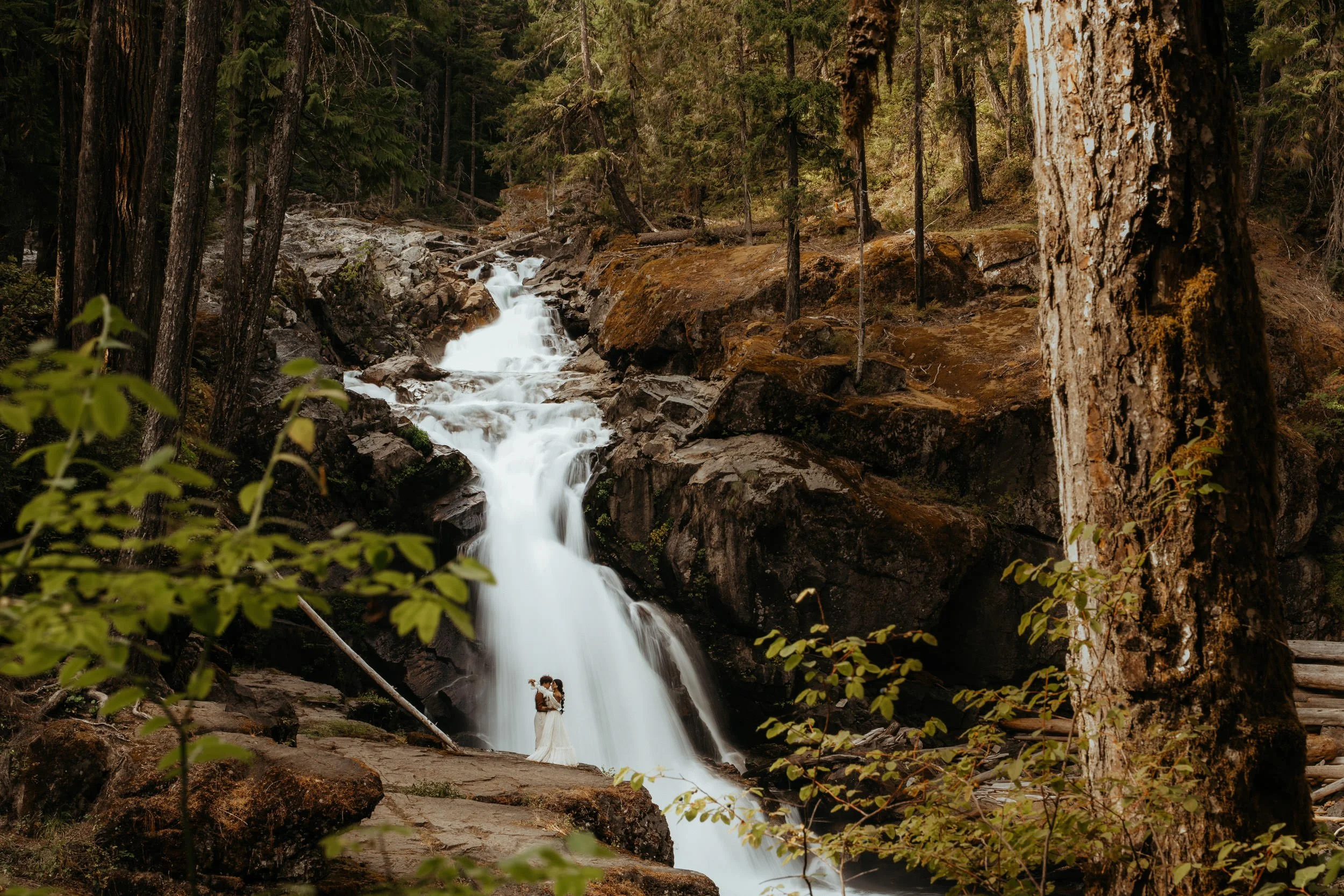 A couple standing close together in wedding attire at the base of a waterfall in a forested area.