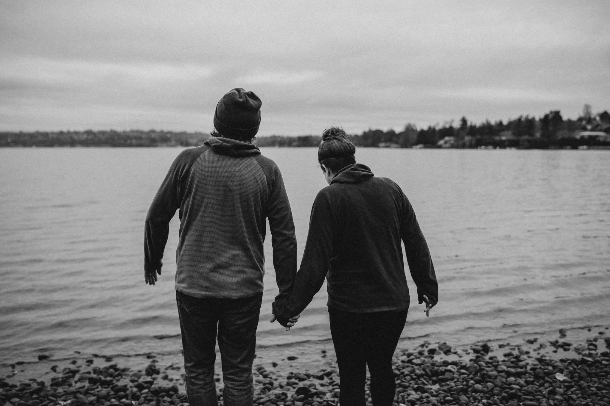 A black and white photo of a couple holding hands and walking along the rocky shoreline by a body of water, with houses visible in the distance.