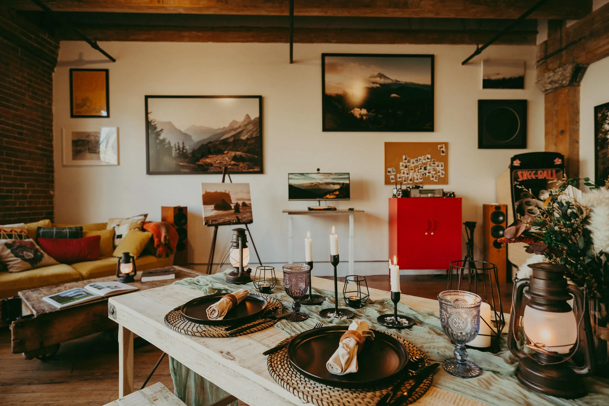 A cozy living room and dining area with a rustic style. The dining table is set with black plates, wrapped napkins, and candles. The living room has a yellow sofa with colorful cushions, and the wall above features framed landscape photographs. There