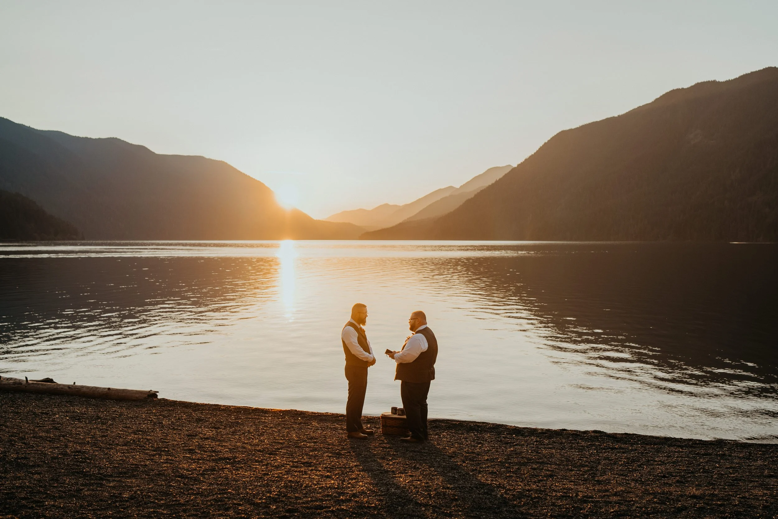 Two men in formal attire standing by a lake at sunset, with mountains in the background, engaging in conversation.