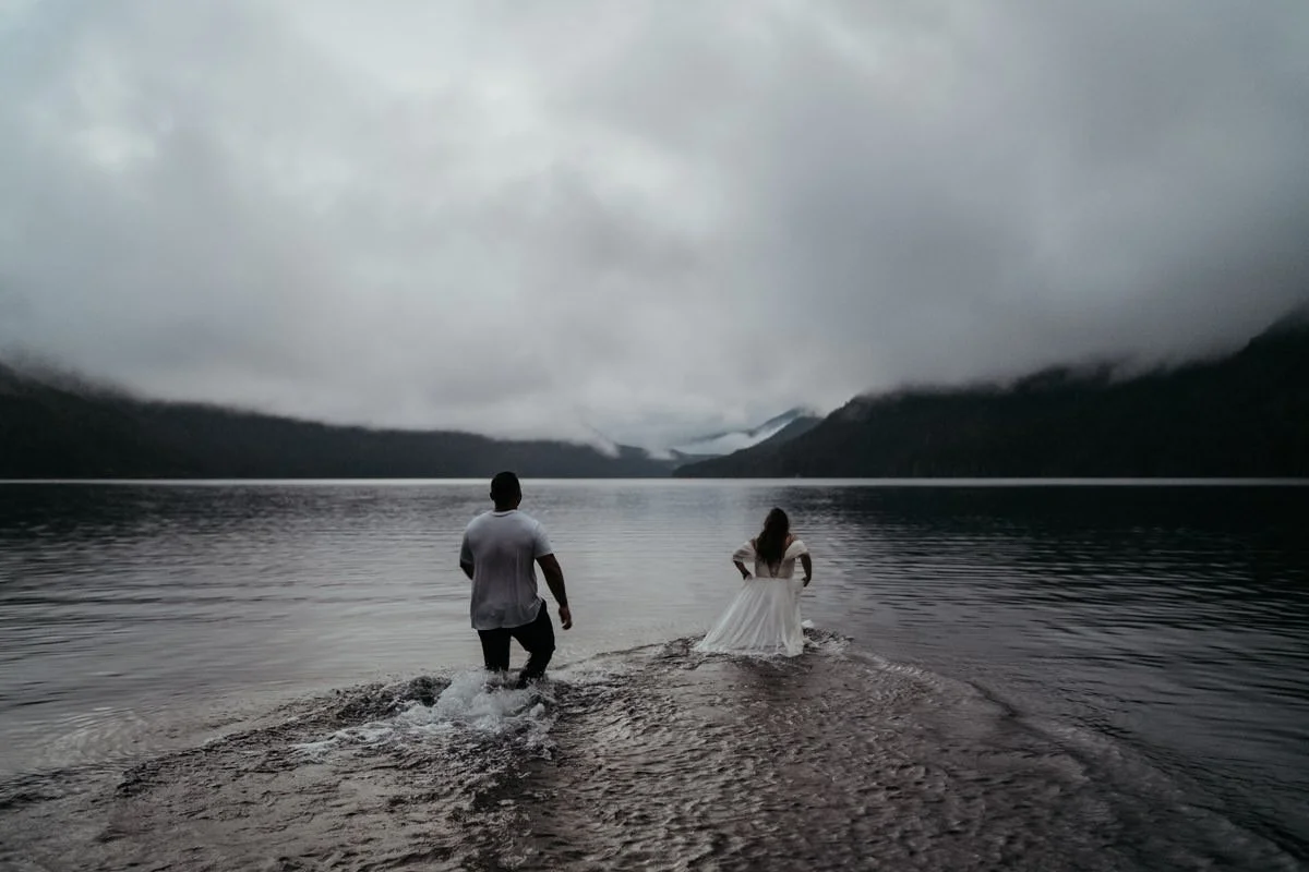 A couple stands in a lake with their backs to the camera, surrounded by mountains and a cloudy sky.