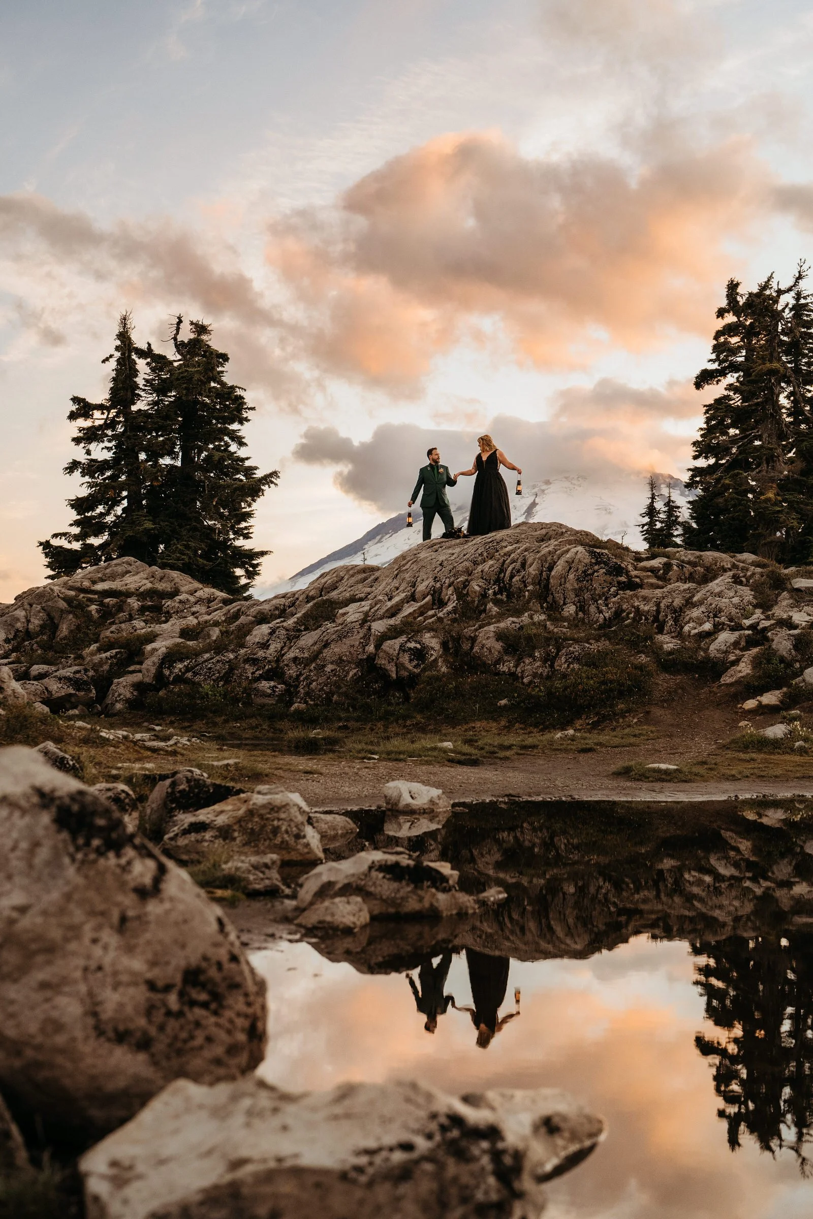 A couple in formal attire standing on a rocky hilltop during sunset, holding hands and drinking beer with a mountain, trees, and a sky with clouds in the background.