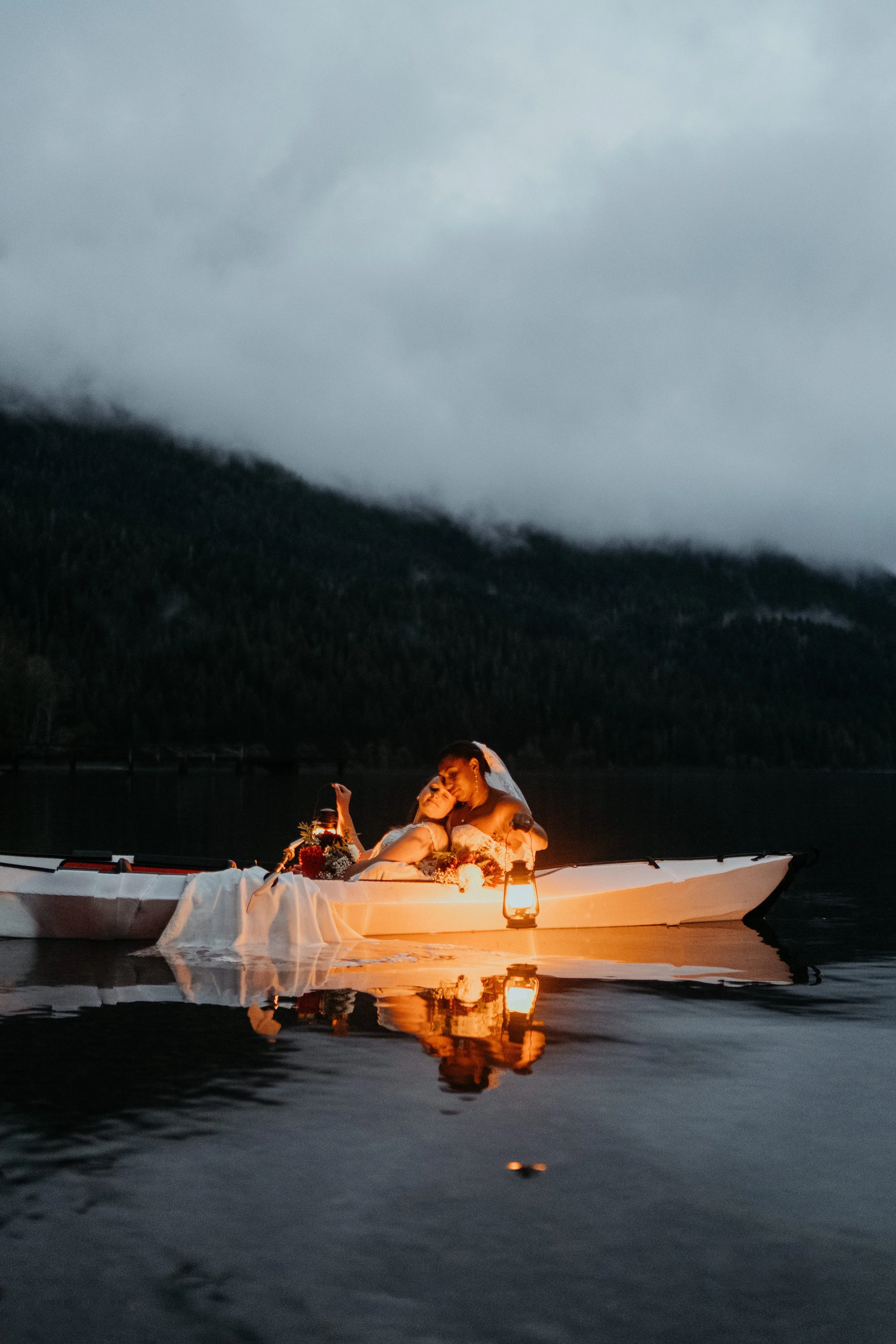 Two women in wedding dresses sharing a kiss in a kayak on a calm lake, illuminated by lanterns, with dark mountains and cloudy sky in the background.