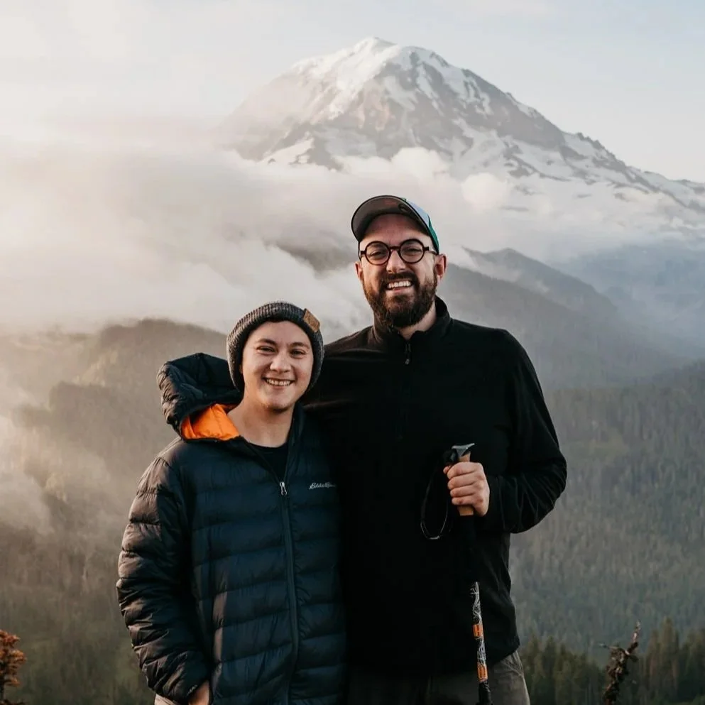 Ashton and Dale, two photographers, standing outdoors with a snow-capped mountain in the background, dressed in outdoor jackets and a beanie.