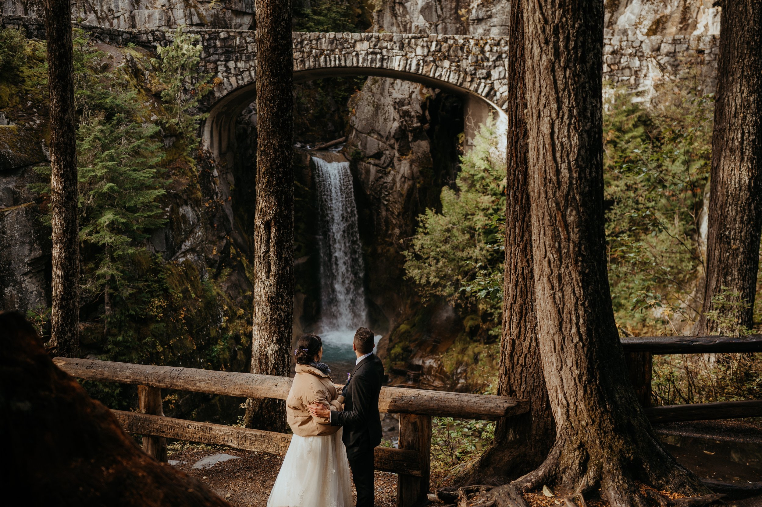 A bride and groom standing near a wooden fence in a forest with tall trees, overlooking a waterfall and rock formation with a stone bridge overhead.