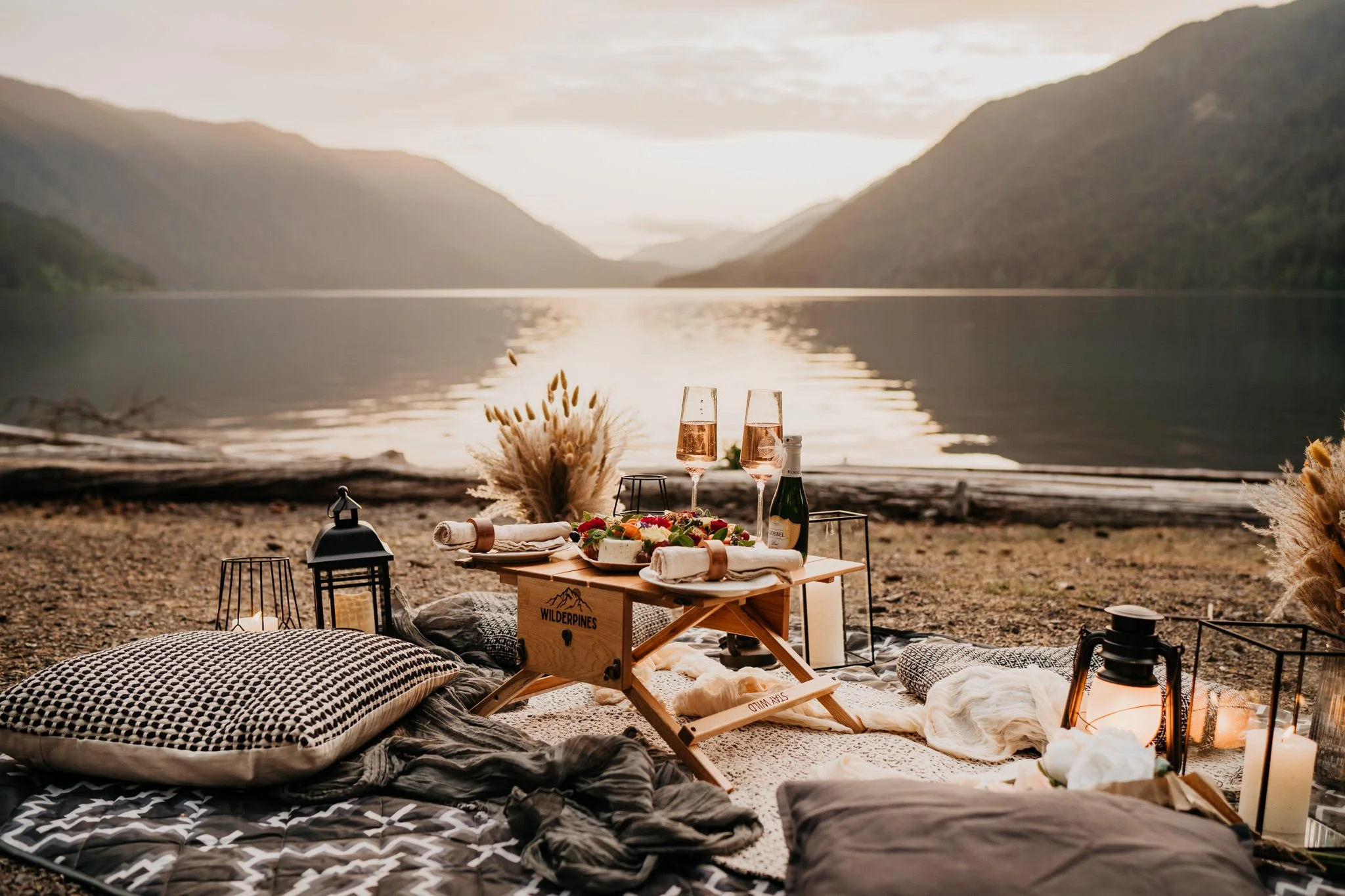 A lakeside picnic setup at sunset with pillows, lanterns, a small wooden table with plates of food and drinks, surrounded by mountains and calm water.