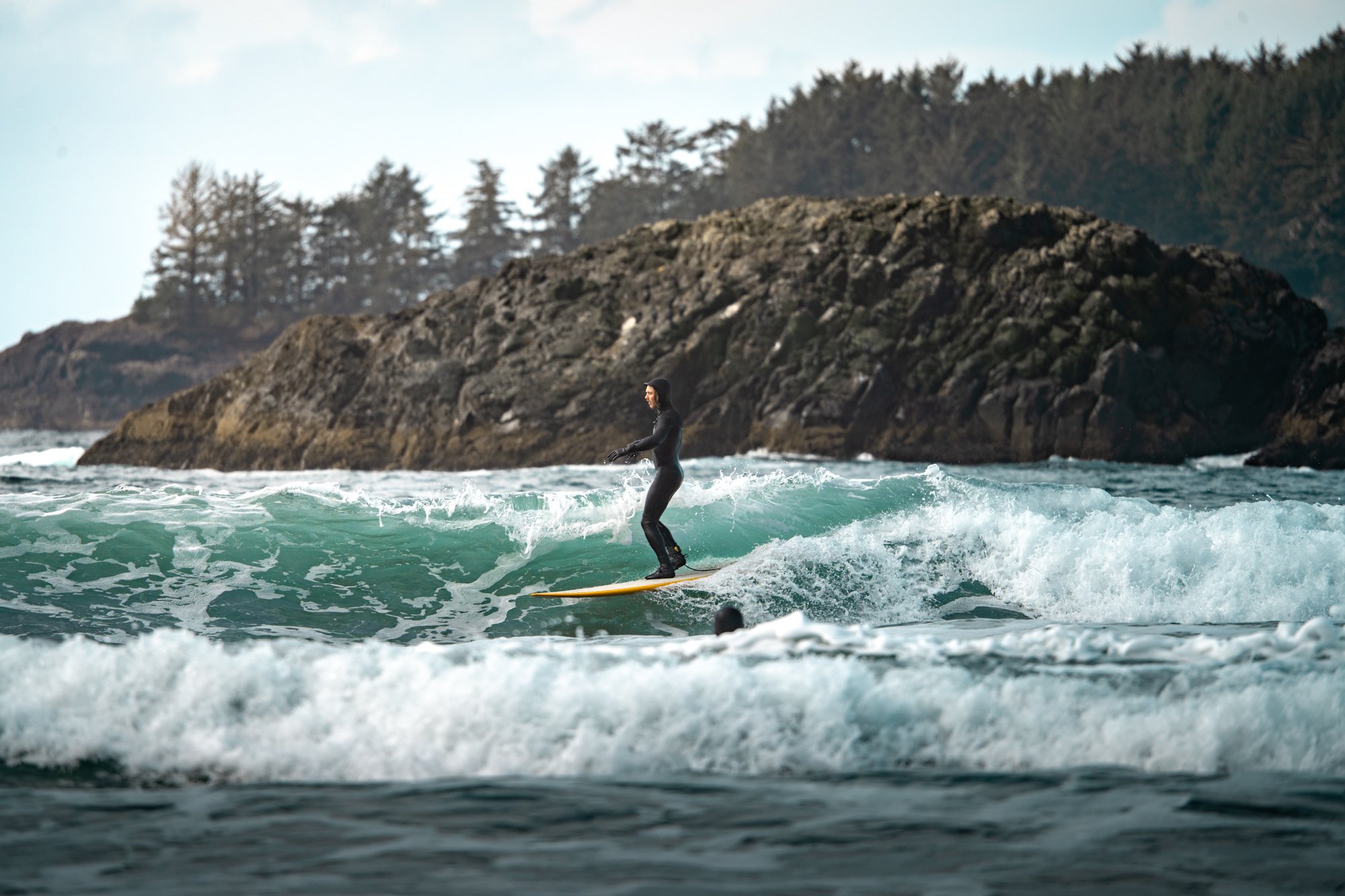 A person wearing a wetsuit surfing on a yellow surfboard in the ocean near a rocky, forested coastline.