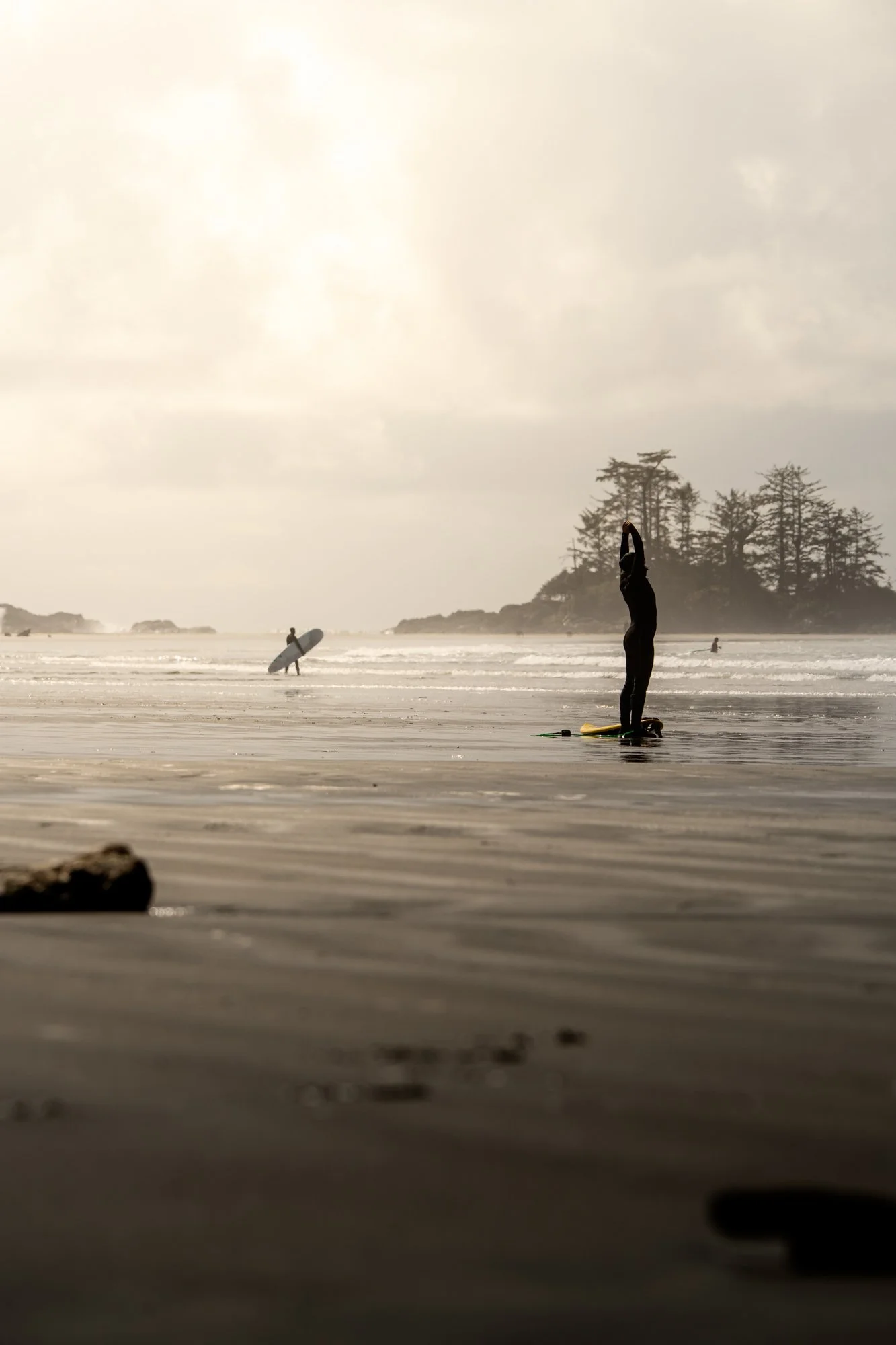 Person standing on a beach by the ocean, with others surfing in the background and an island with trees in the distance in Tofino, British Columbia.
