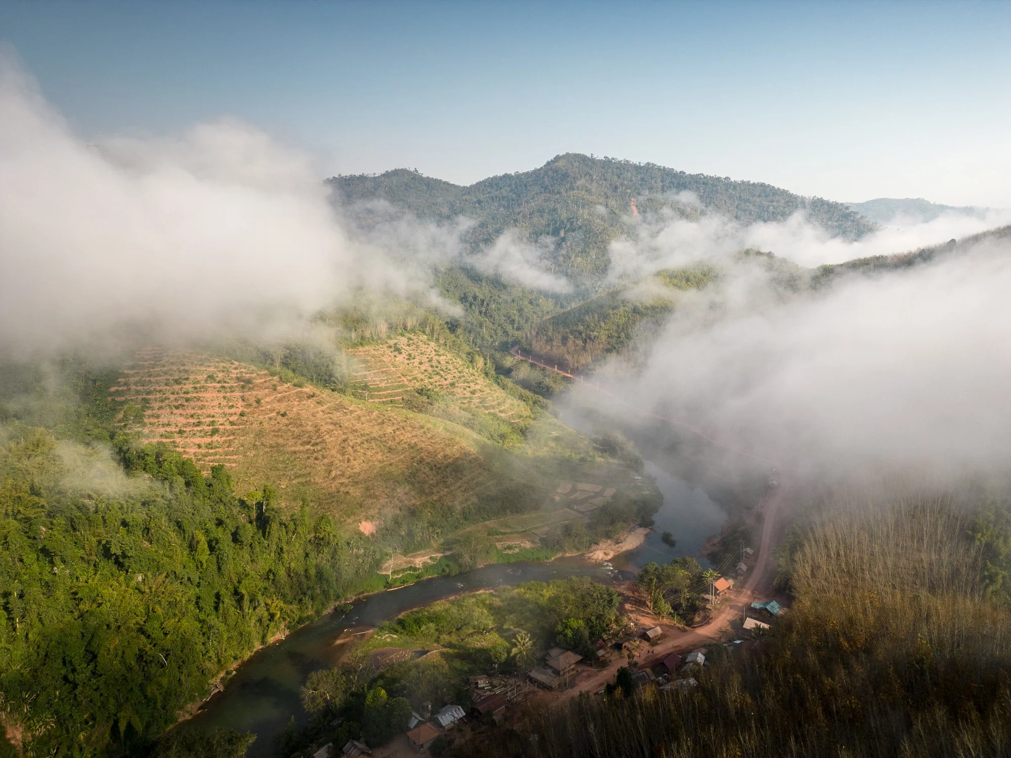 Aerial view of a river flowing through a lush green valley surrounded by mountains with fog creeping over the landscape, Laos.