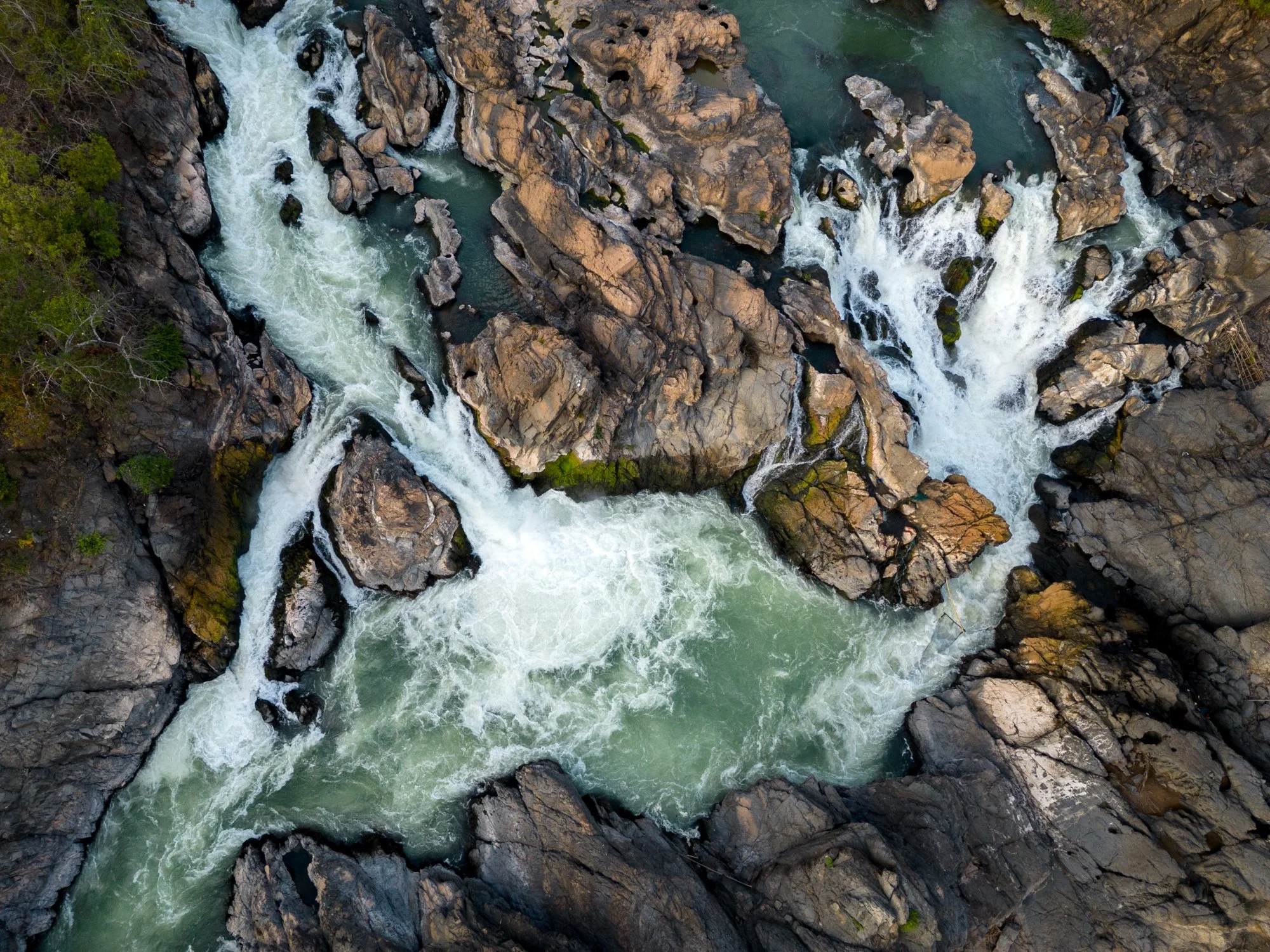 Aerial view of a fast-moving river with white water rapids flowing through rocky terrain surrounded by greenery on the Mekong River, in Laos.