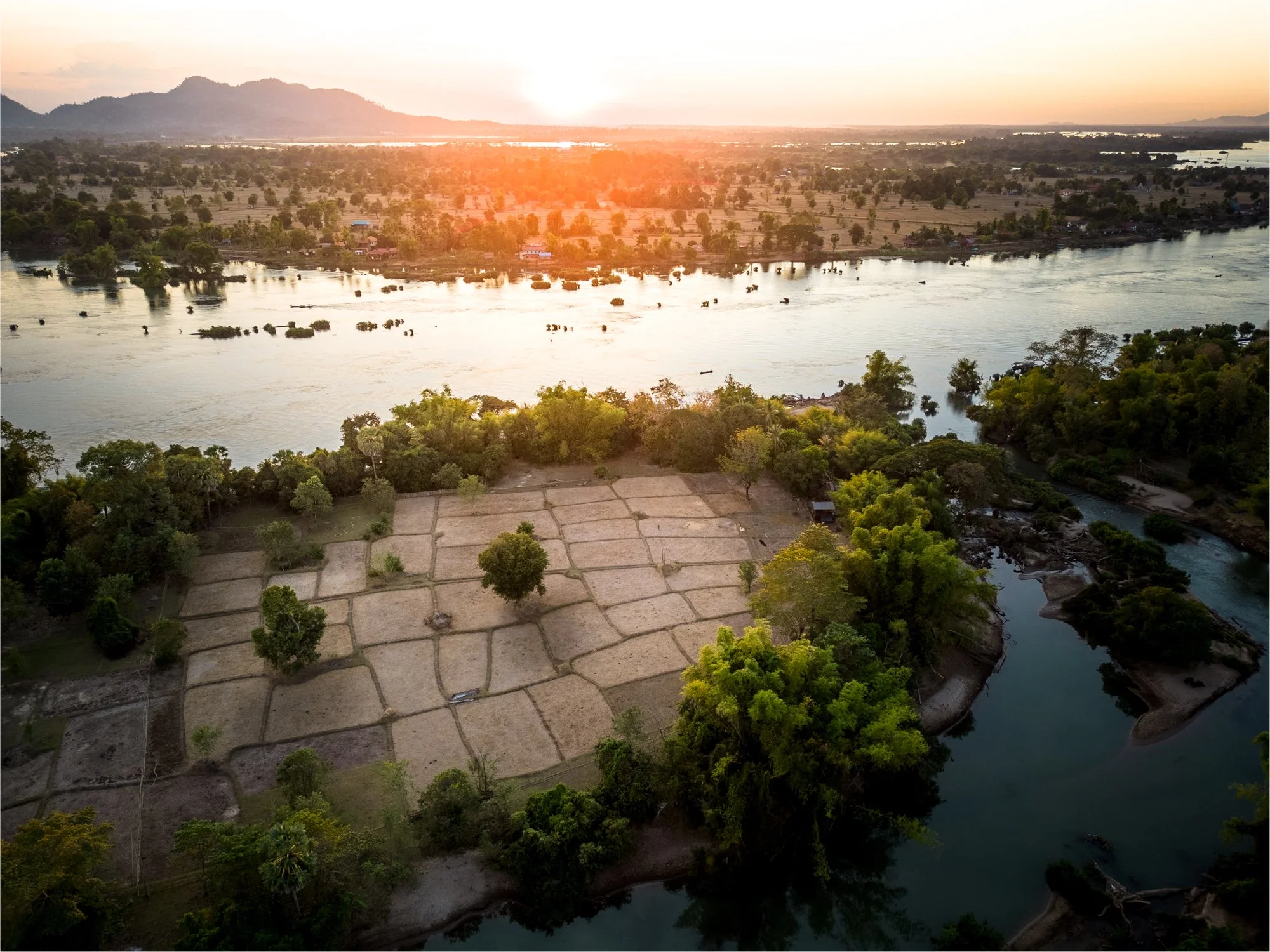 Aerial view of the Mekong river flowing through lush green Laos landscape with farmland and trees, with a sunset or sunrise casting a warm glow over the scene.