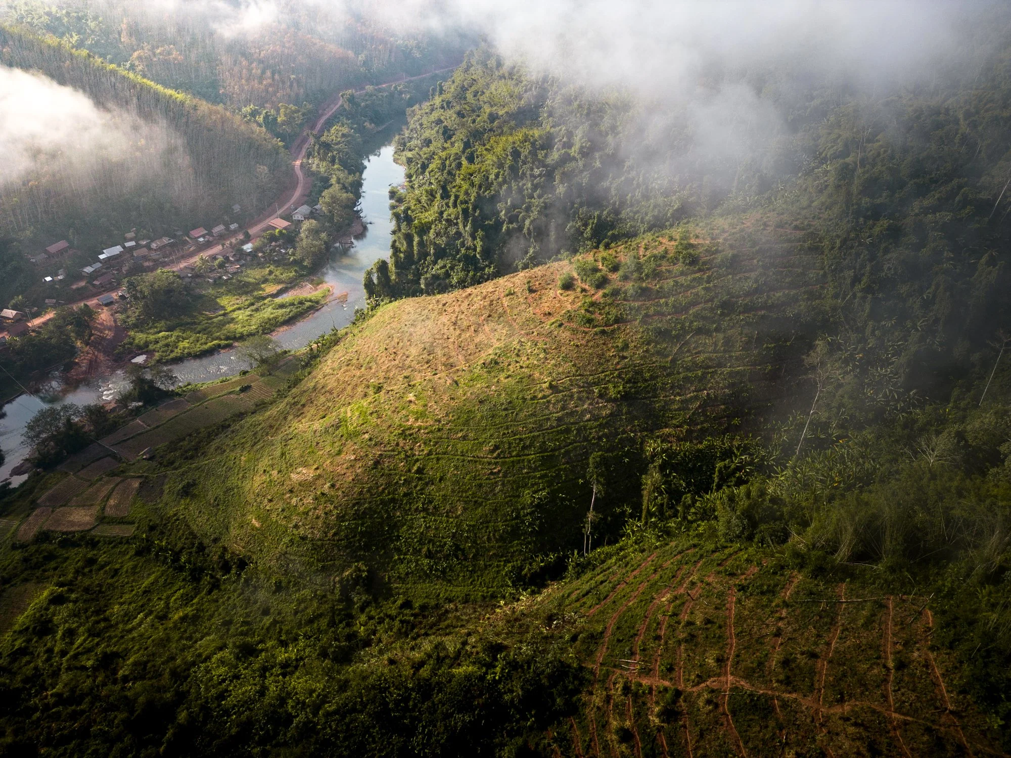 Aerial view of a lush green mountain in Laos with terraced fields, a river, and a village in a misty valley.