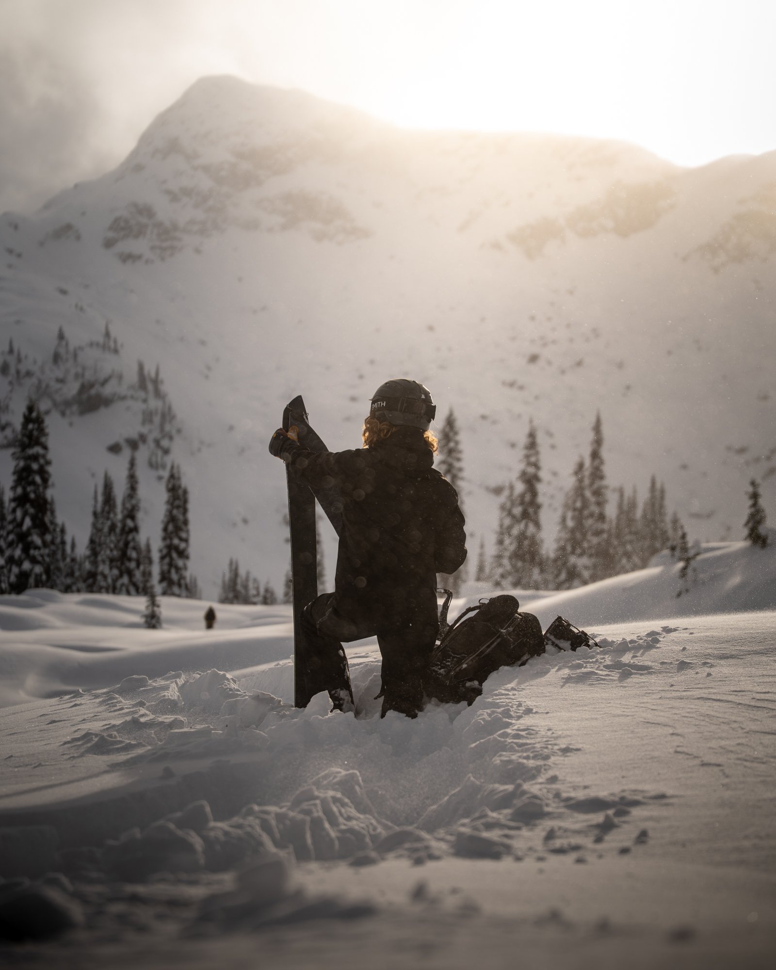 A person in black winter gear, wearing a helmet, kneeling in deep snow and holding a snowboard, with a snowy mountain and pine trees in the background, during sunset at the Wendy Thompson Hut near, Pemberton, British Columbia.