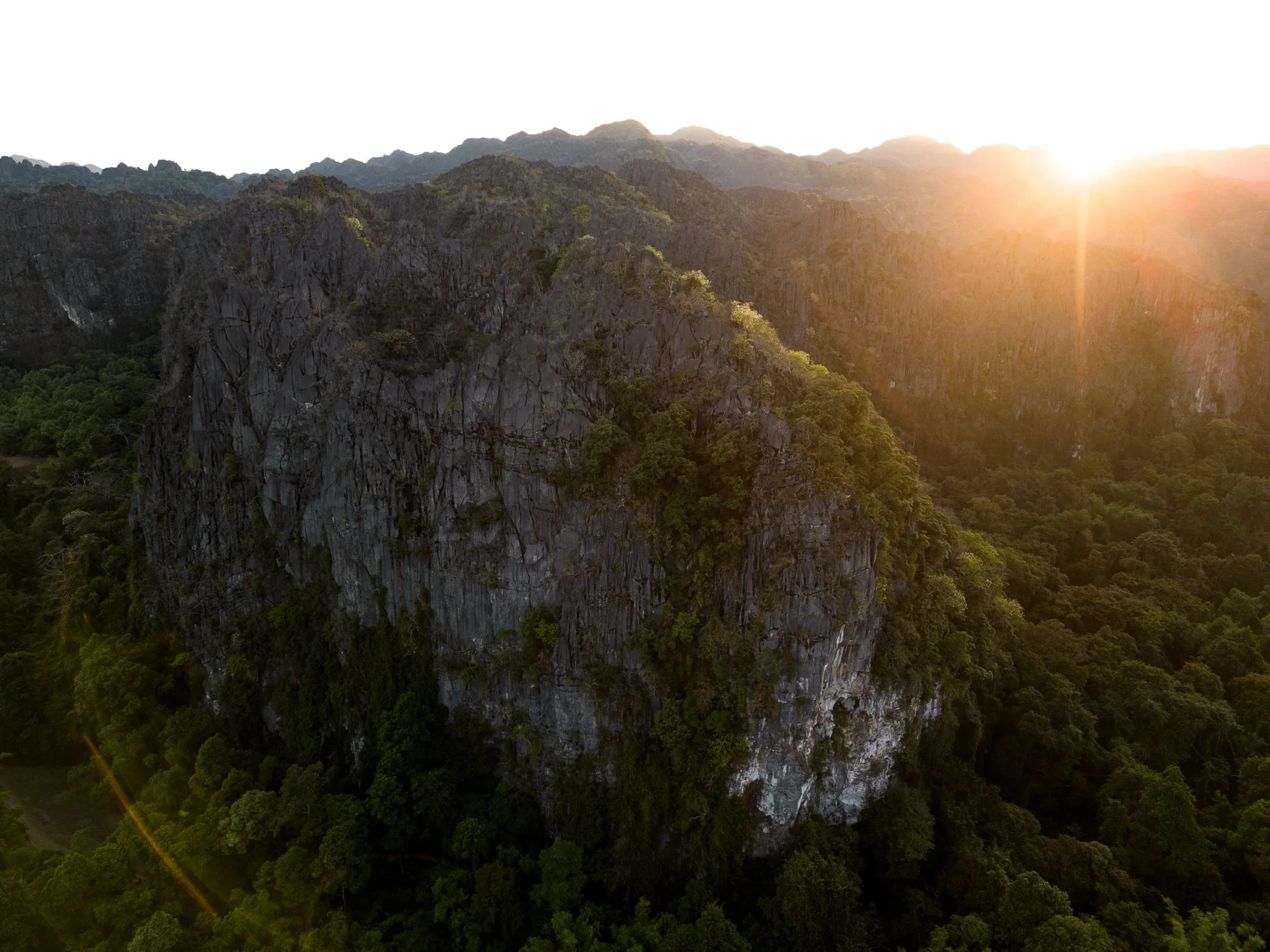 Aerial view of a large rocky mountain in Laos with dense green forest at its base, illuminated by the setting or rising sun.