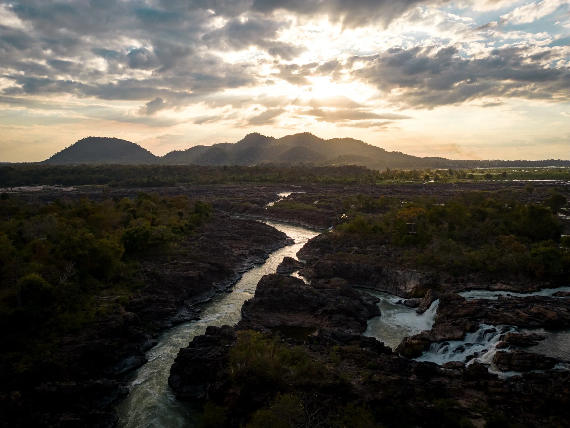 Sunset over the Mekong river flowing through the rocky landscape of Laos, with mountains in the background and partly cloudy sky at sunset.