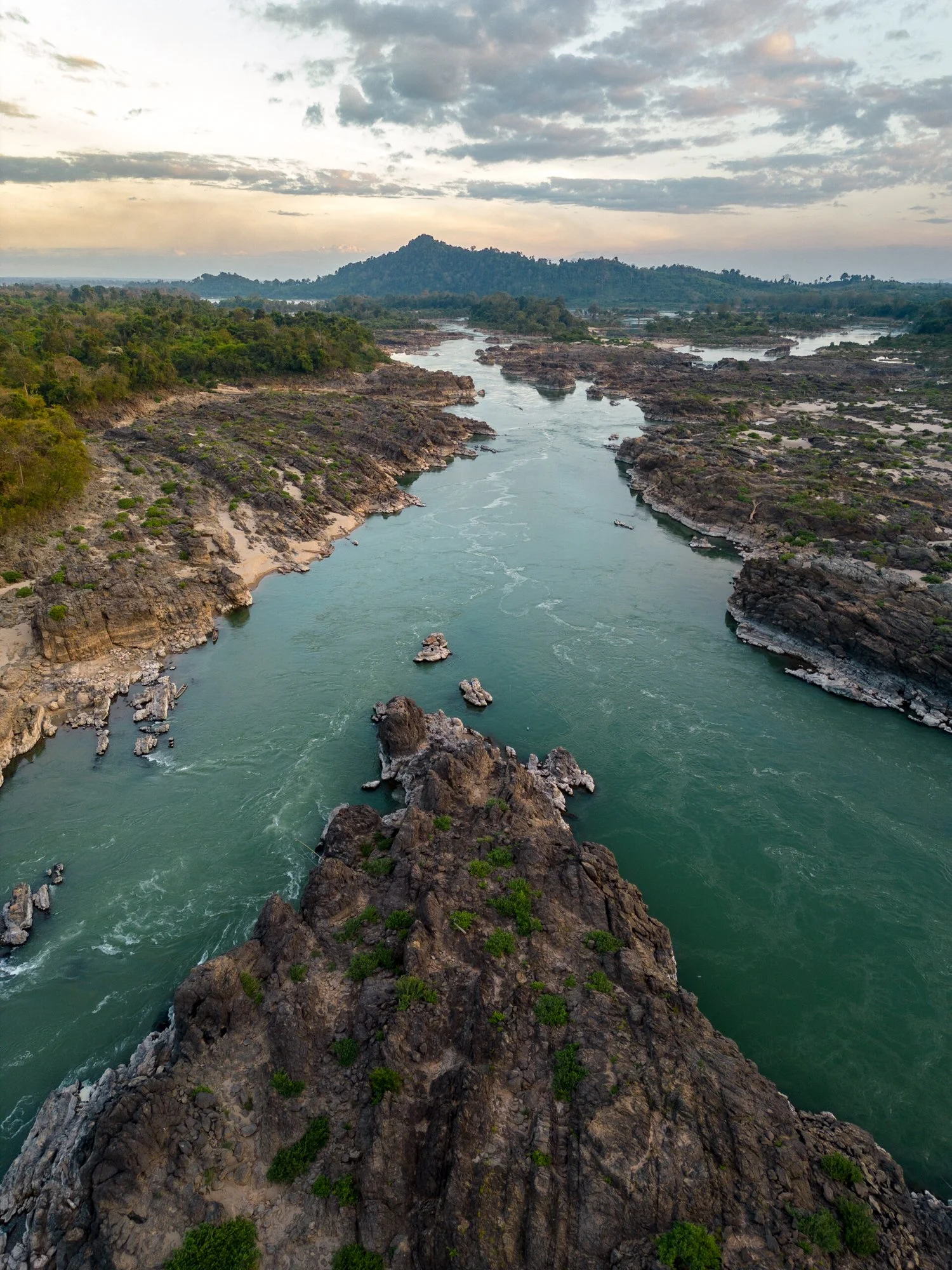 An aerial view of  the Mekong river flowing through rocky and forested terrain of Laos, with a mountain in the background under a cloudy sky at sunset.