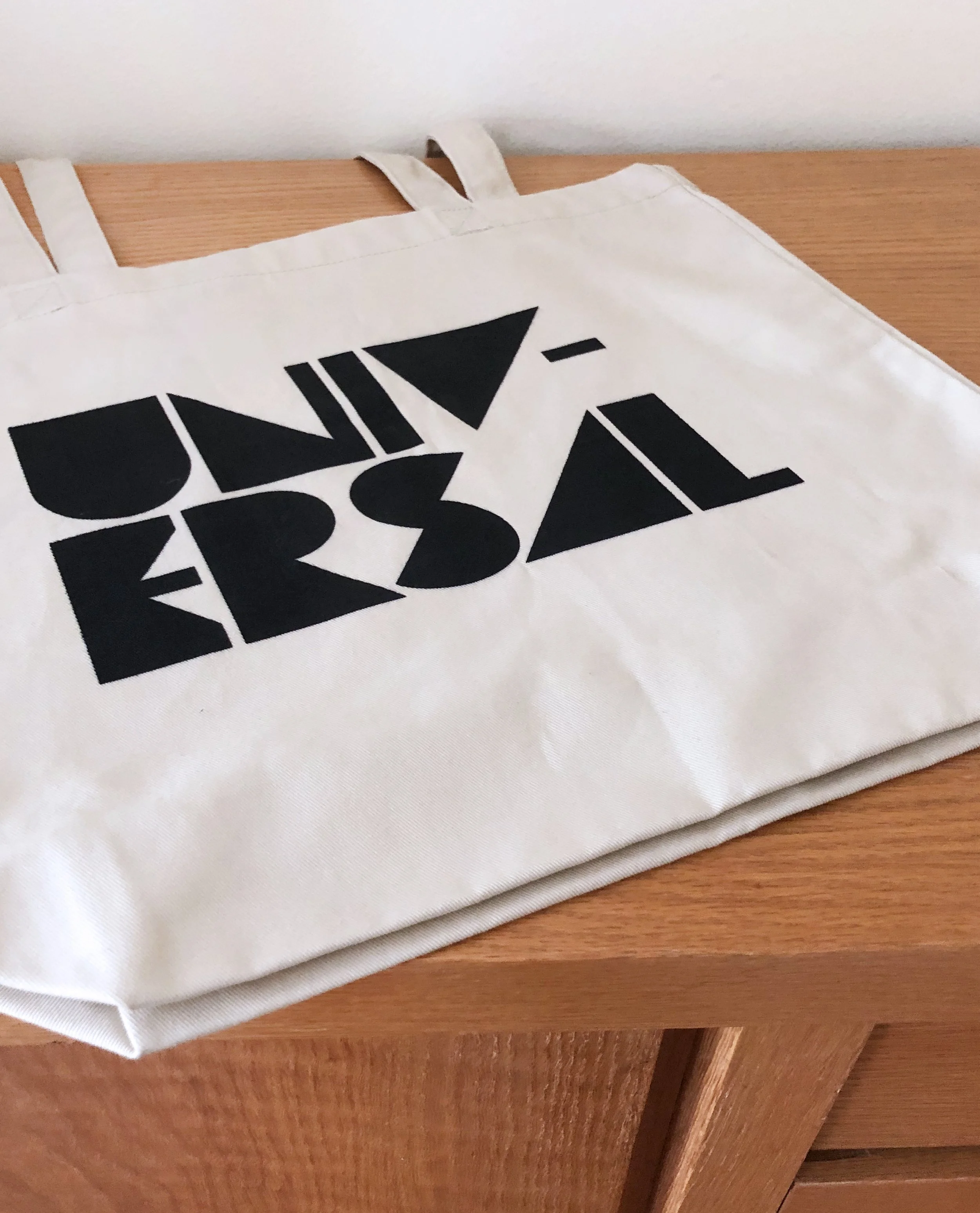 Image of a beige tote bag sitting on top of a modern, wooden dresser. The word, Universal, is printed in black in a block font on the front of the tote.