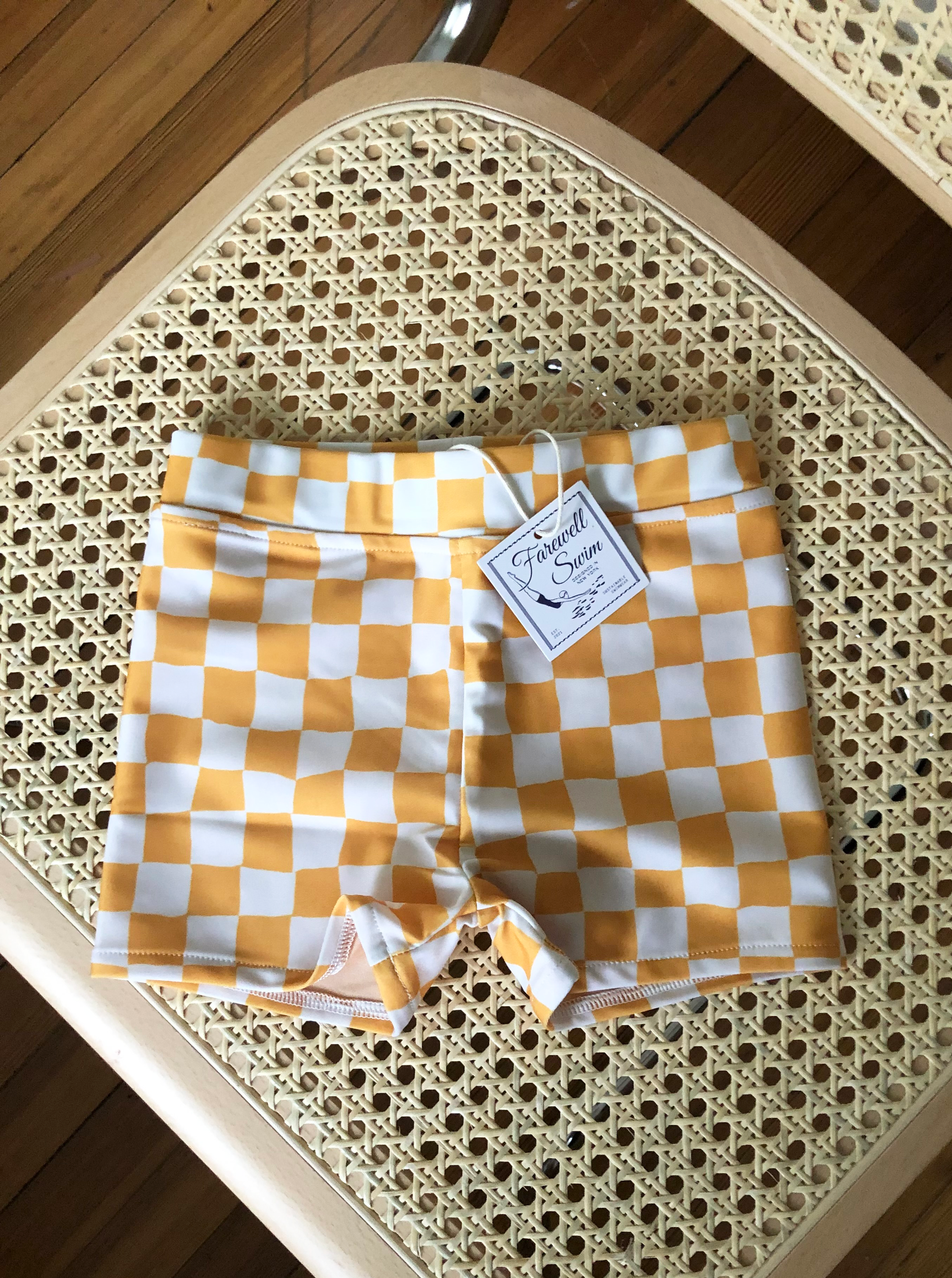 Photo of kids yellow and white checkered swim trunks on a cane chair with a Farewell Swim tag hanging out of the front of them.