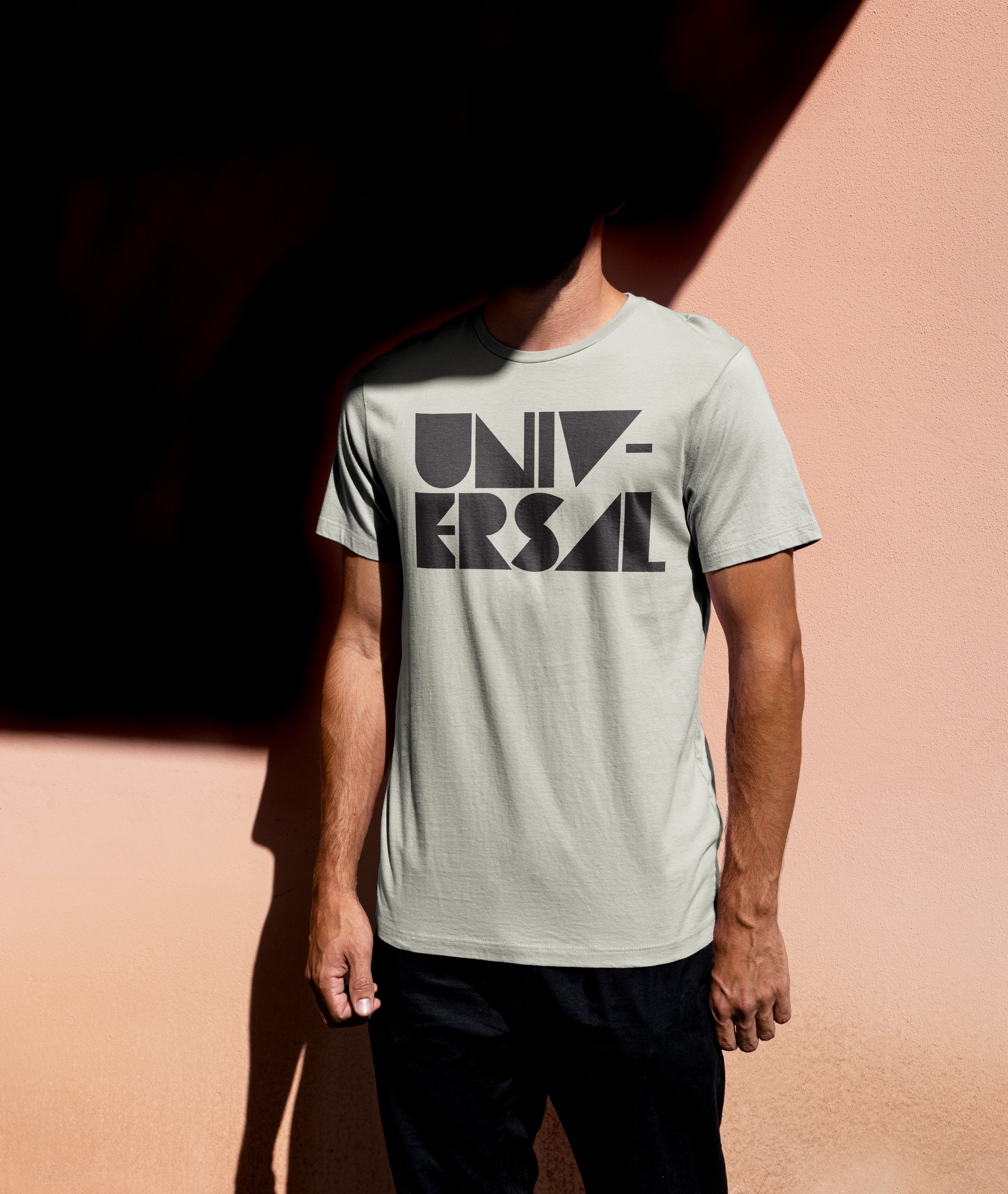 Image of a man in front of a pink wall with a shadow over his face and a beige t-shirt with the word, "Universal" in black. He is also wearing black jeans.