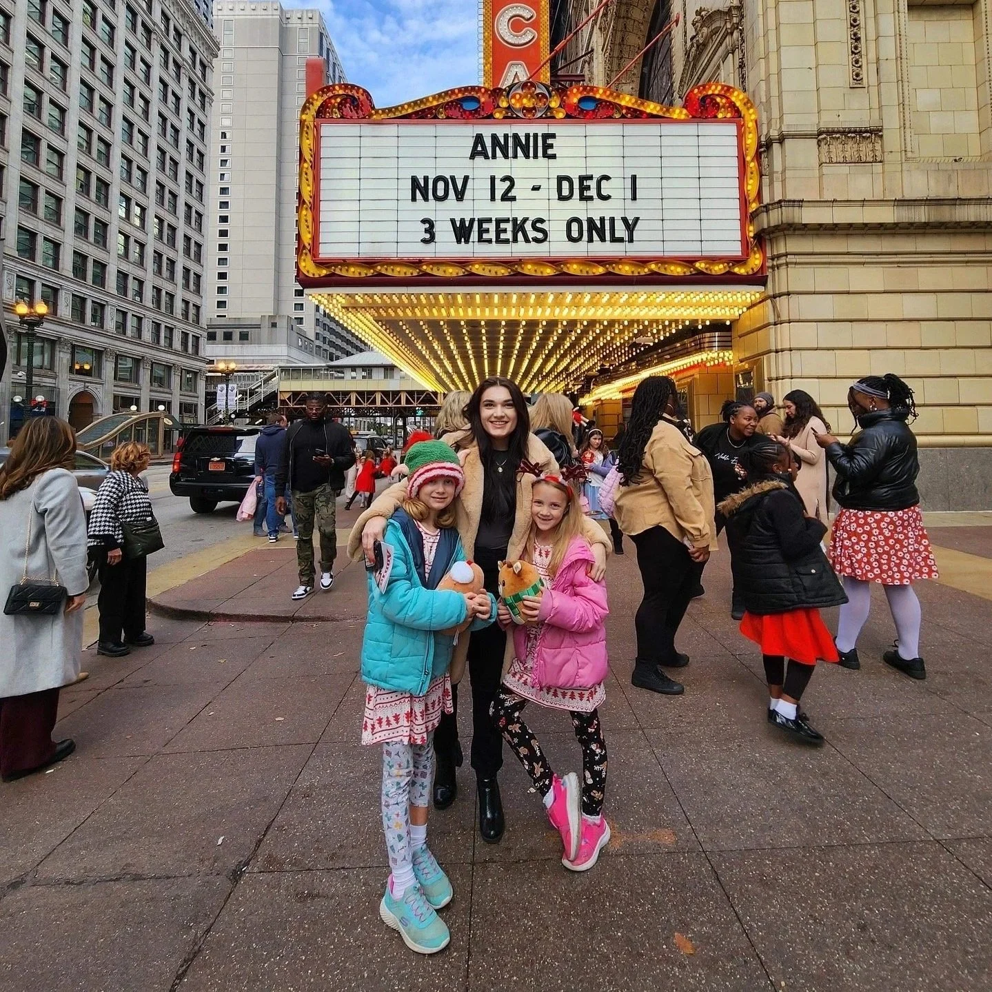 People standing in front of a marquee that advertises a show, with a group of children and an adult smiling at the camera. The marquee reads "ANNE NOV 12 - DEC 1 3 WEEKS ONLY." The scene appears to be outside a theater, with tall buildings and a busy pedestrian area.