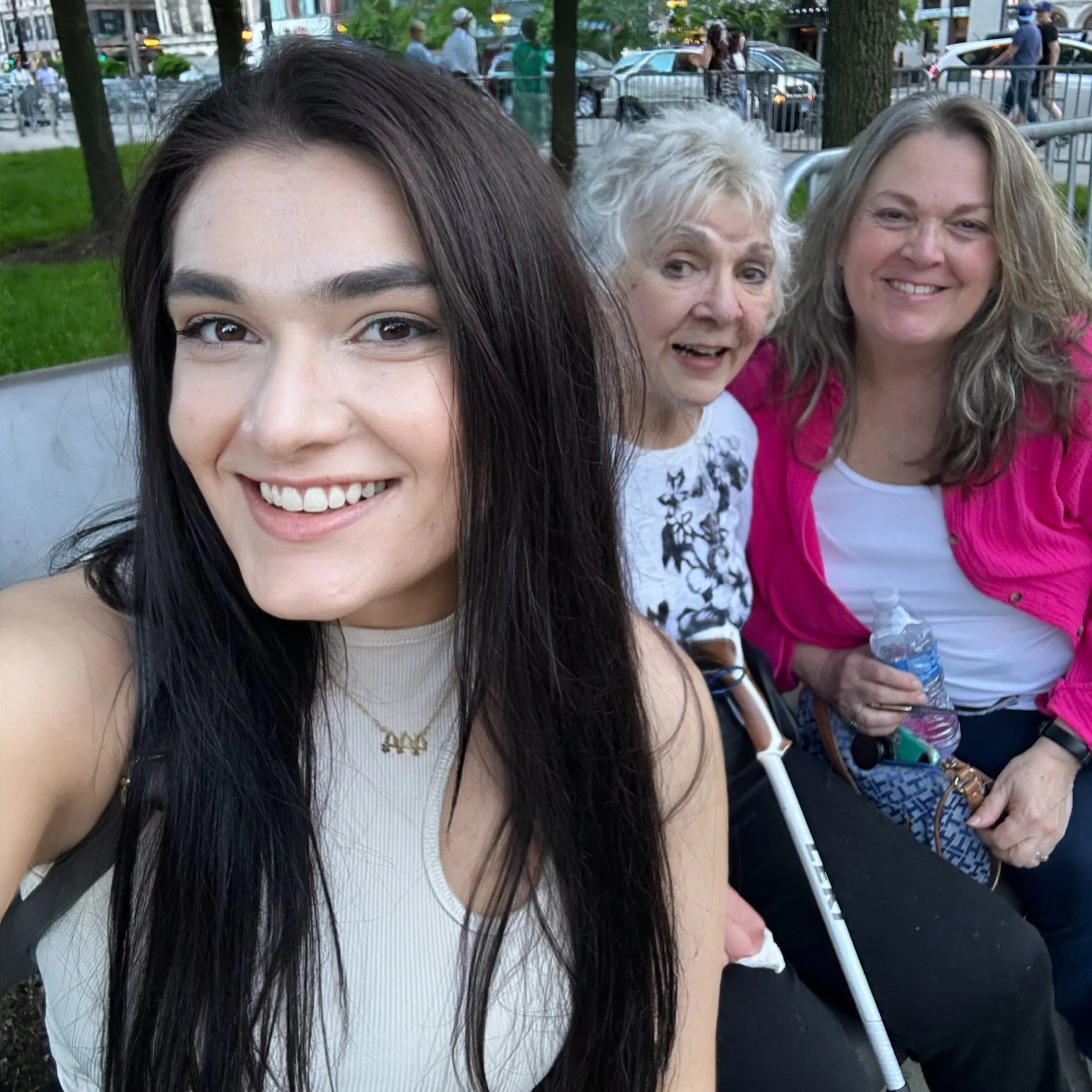 Close-up of three women sitting outdoors in an urban park, with trees and parked cars in the background, taking a selfie.