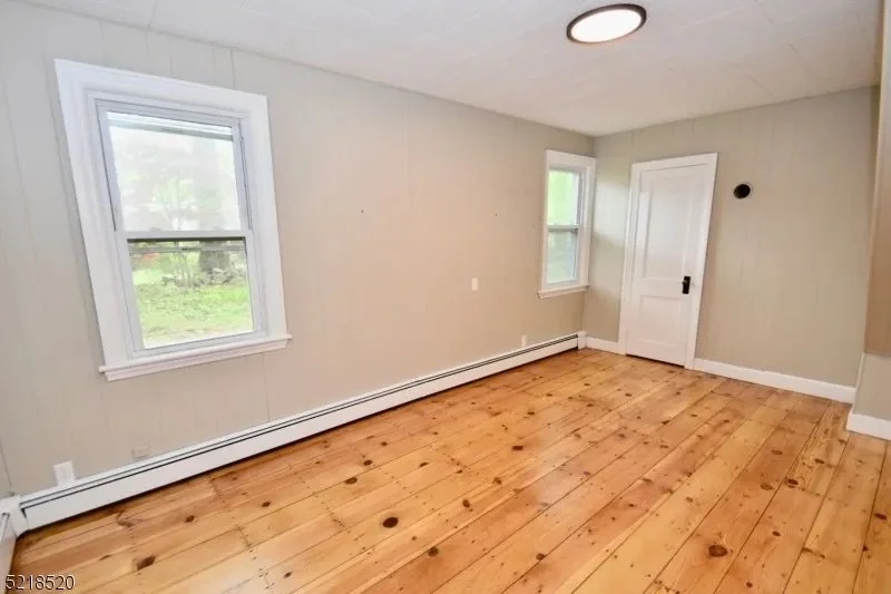 An empty room with beige walls, two windows, a white door, and wooden plank flooring.