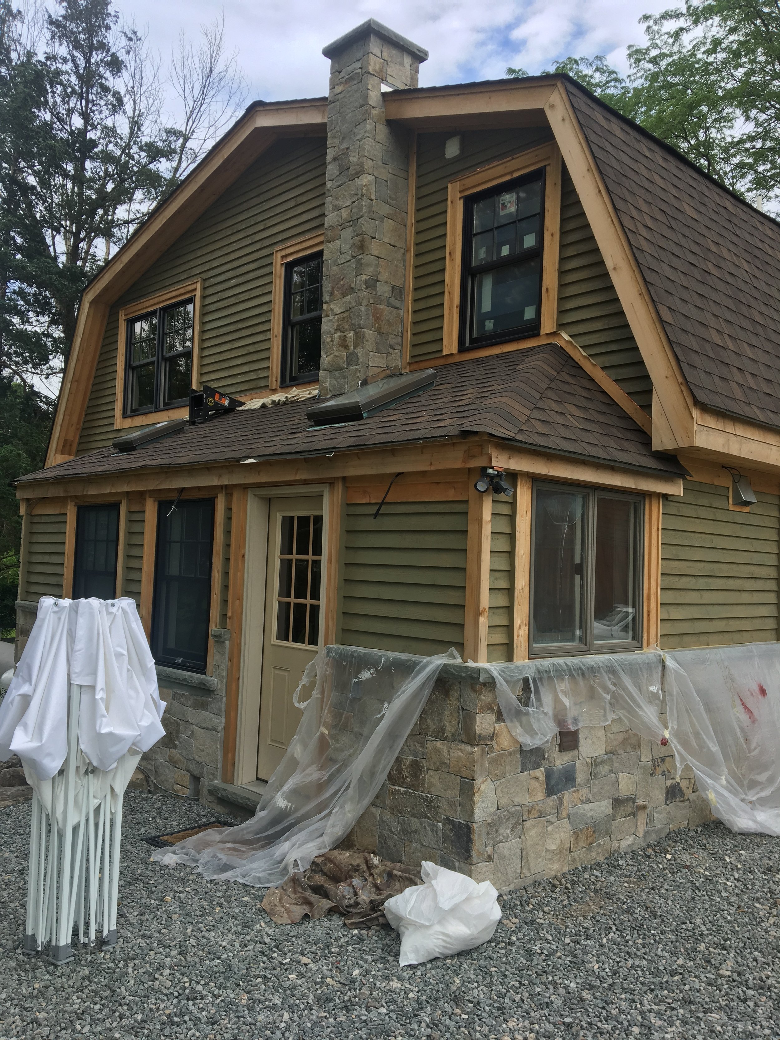A two-story house under construction with a stone foundation, green siding, and a steep brown shingle roof. The house features multiple black-framed windows and a chimney. Protective plastic sheeting is present, indicating ongoing work.