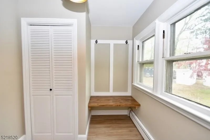 A small room with beige walls, a white louvered closet door on the left, two windows on the right, a wooden bench with a back wall panel between the windows, and a baseboard heater underneath the windows.