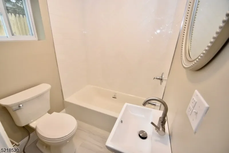 Small bathroom with a window, a toilet, a bathtub, a small sink with a modern faucet, a round mirror, and a wall switch.