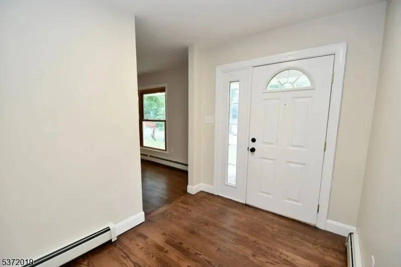 Entryway with a white door with an arched window and side windows, hardwood floor, and a glimpse into a living room with a window and hardwood flooring.