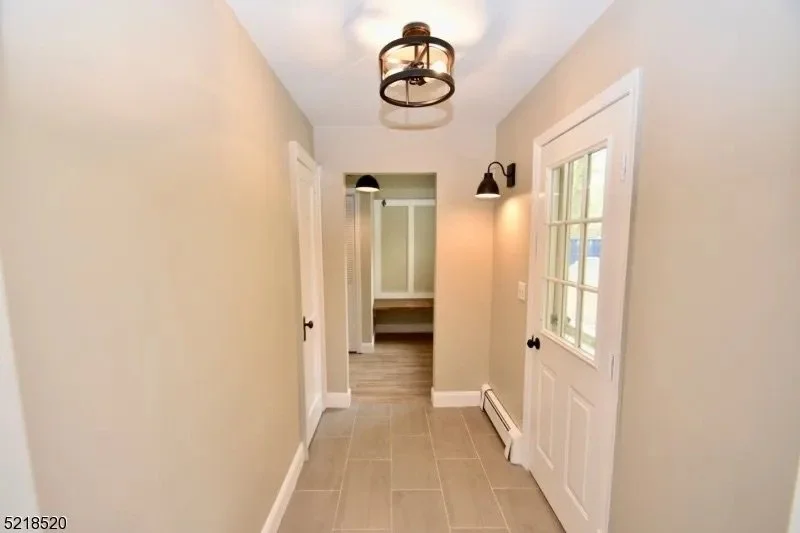An entryway in a house with beige walls, a ceiling light fixture, wall-mounted black lamps, a windowed door, and a view into a hallway with a door and a small sitting area.