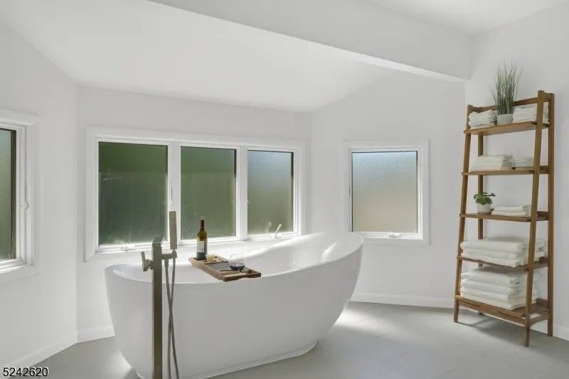 Minimalist bathroom with a white freestanding bathtub near three windows, a wooden ladder shelf with folded towels and plants, and a floor-mounted faucet.