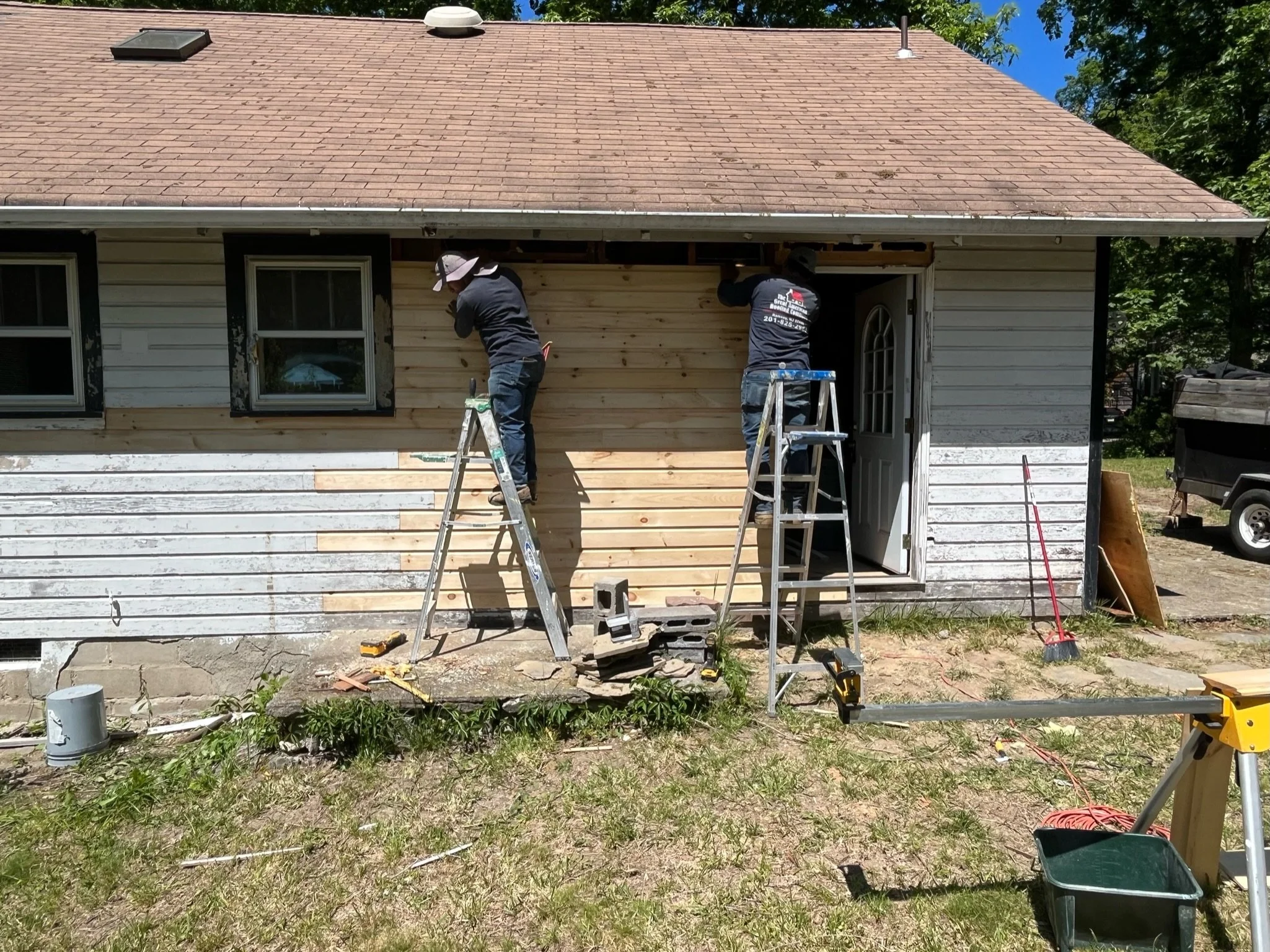 Two workers installing new siding on a house using ladders, with tools and construction materials around them, during daytime.