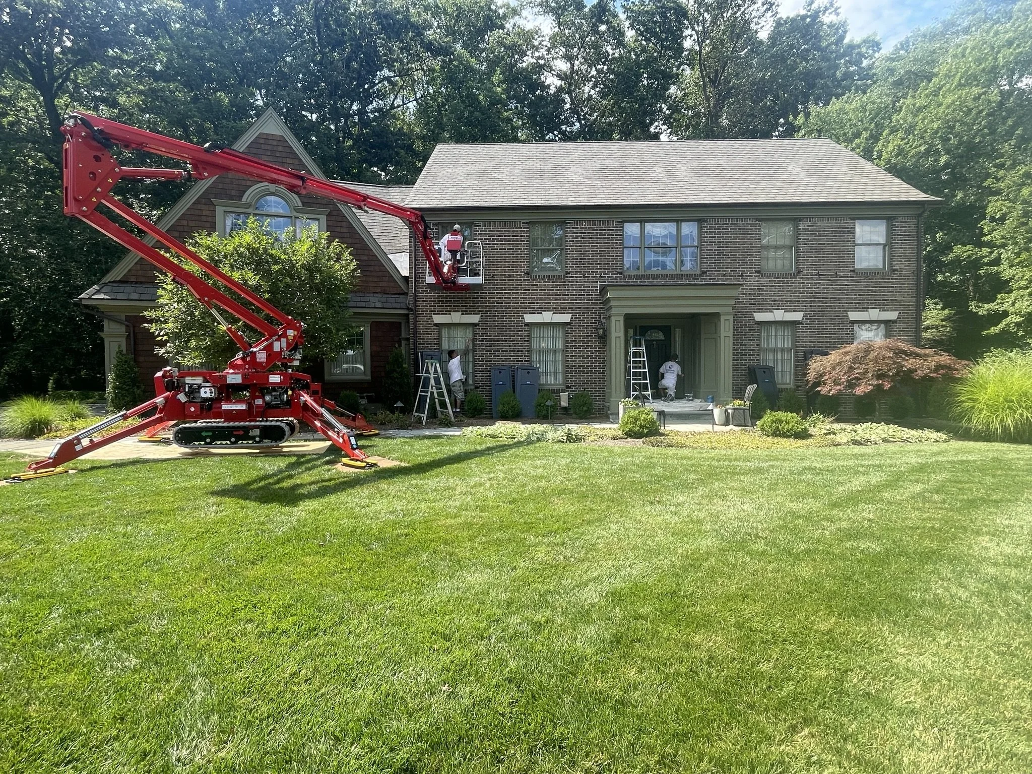 Workers using a cherry picker lift to work on the exterior of a brick house with a front yard and lush green trees in the background.