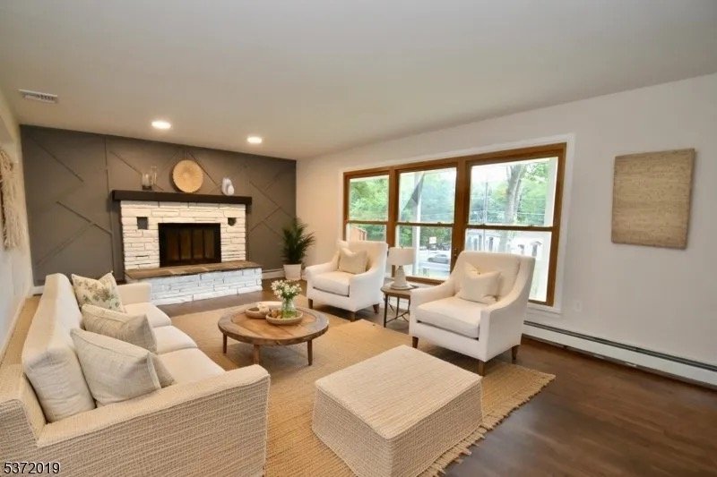 Living room with a beige sofa, two white armchairs, a wooden coffee table, a brick fireplace, large windows, and wooden floors.