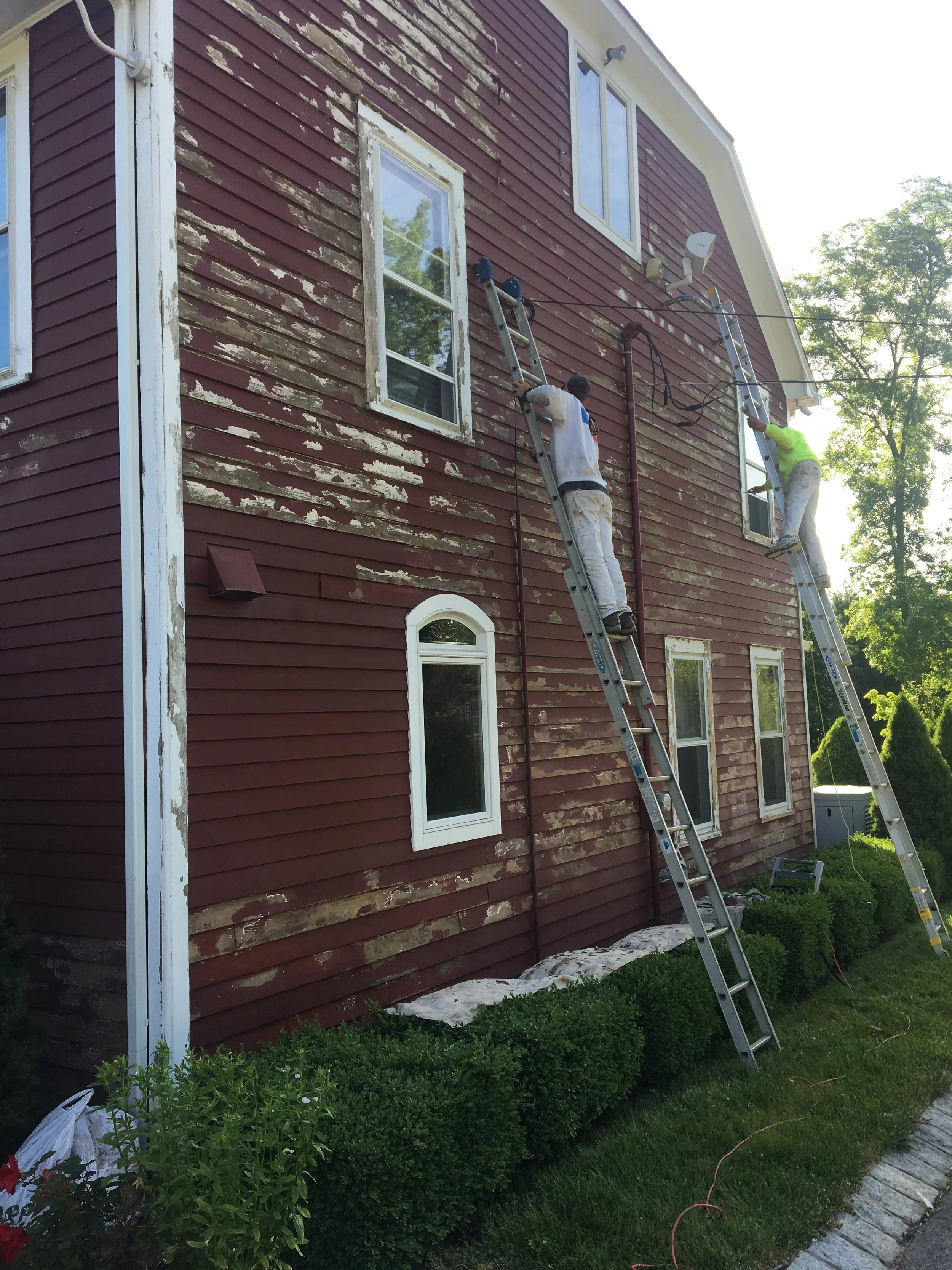Two workers on ladders repairing the exterior of a house with peeling red paint, green bushes in front, and trees in the background.