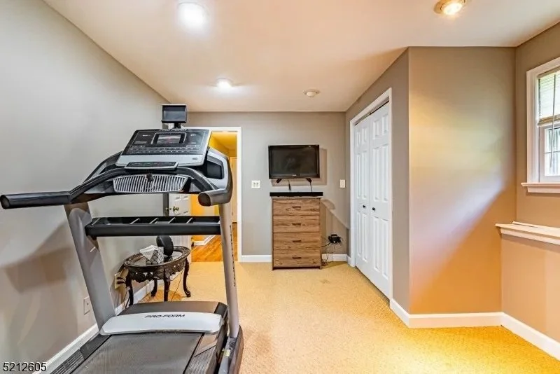 Room with treadmill, small dresser, wall-mounted TV, and windowed wall, beige carpet, and neutral-colored walls.