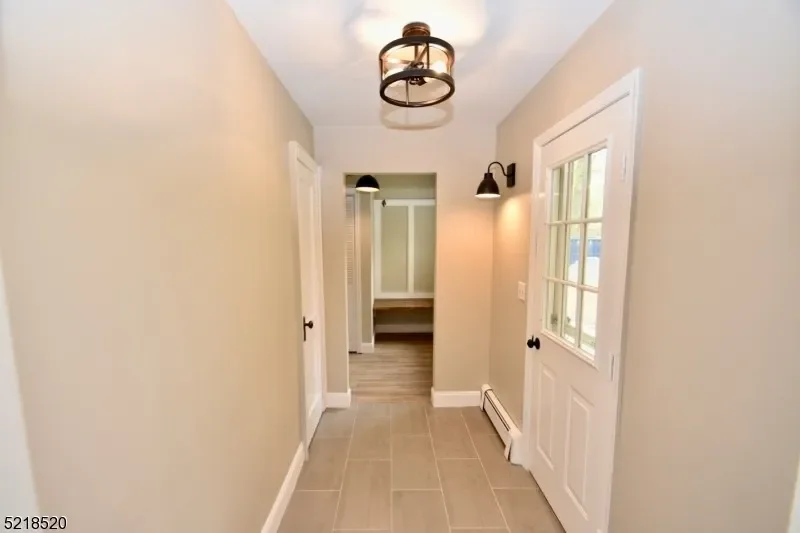 A hallway with beige walls, white door trim, and gray tiled flooring. There are black wall-mounted light fixtures and a ceiling light. At the end of the hallway, there is an open door leading to a room with a wooden floor and white walls.