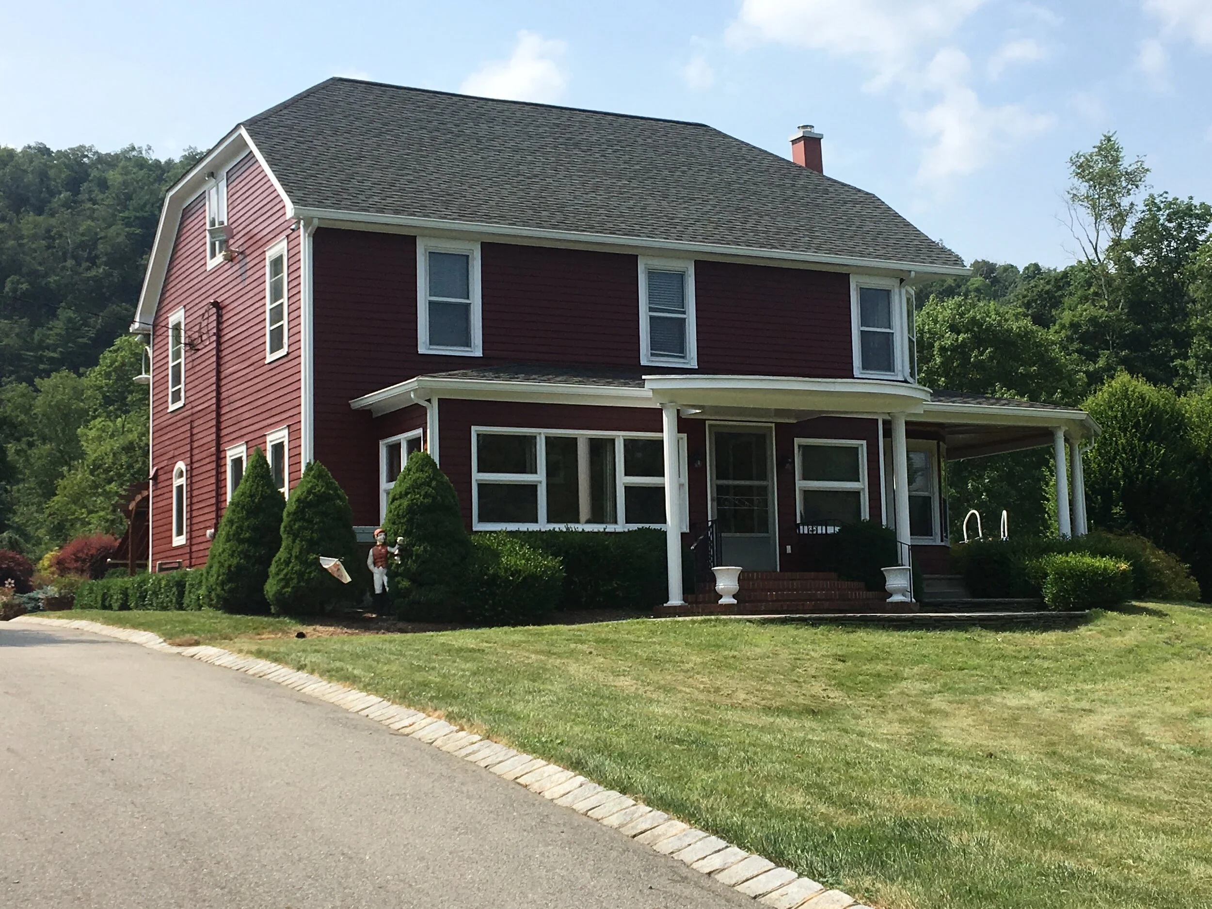 A red, two-story house with white trim, surrounded by green bushes and trees, under a blue sky with some clouds.