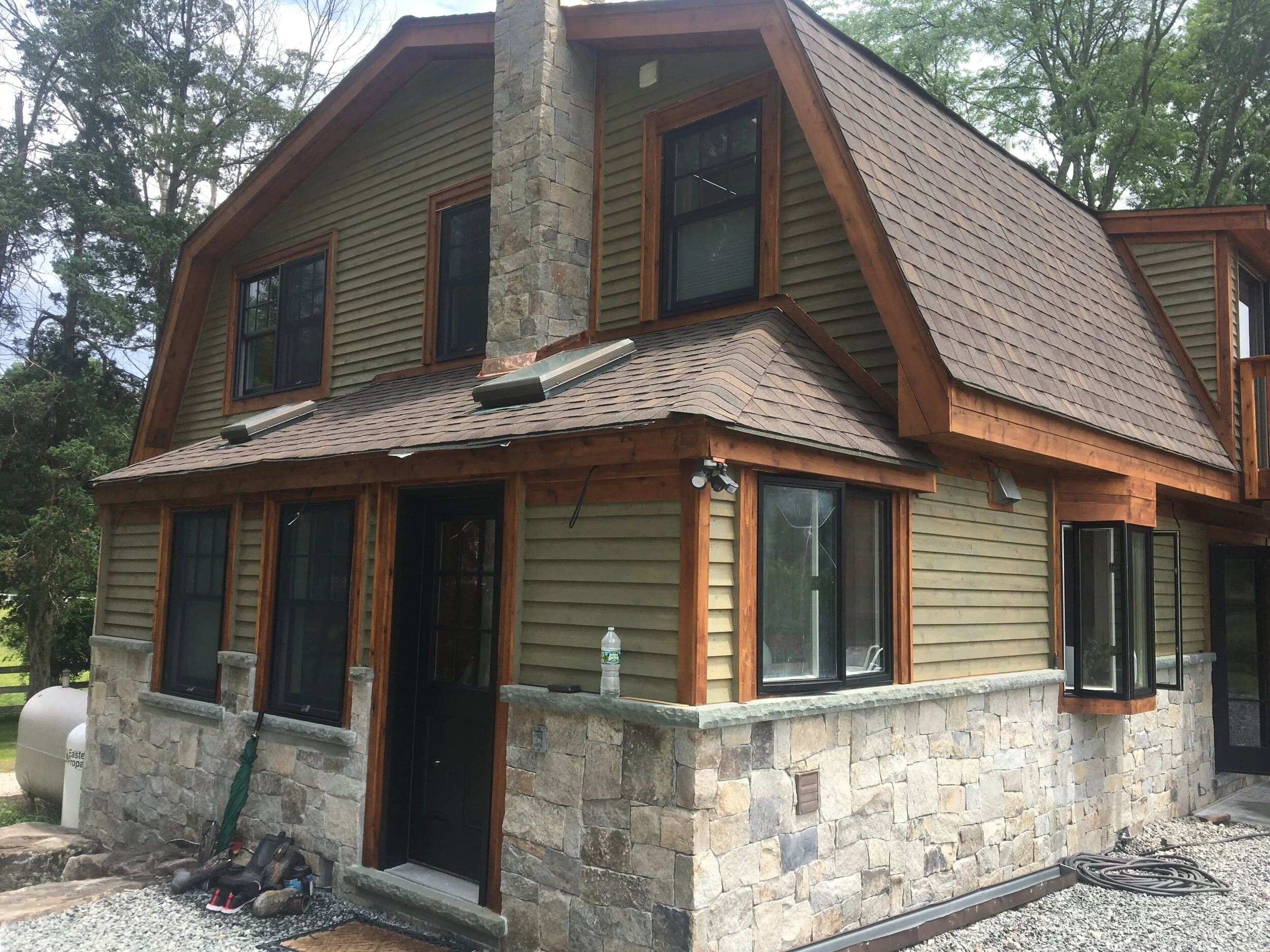 A two-story house with stone and wooden siding, multiple windows, a sloped roof, and a chimney, situated in a wooded area.