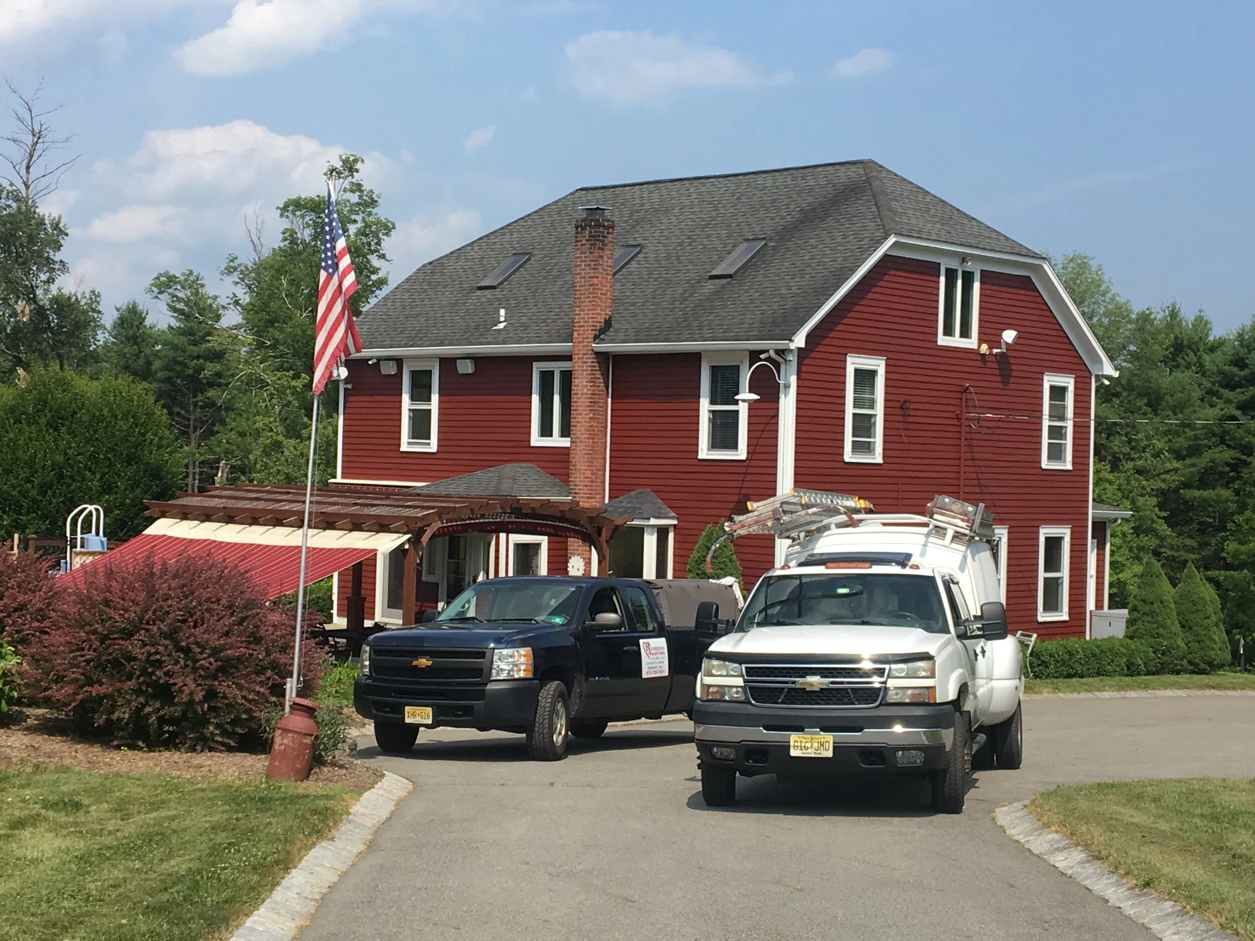 A red house with white trim and numerous windows, two trucks parked in front, a flagpole with an American flag, a shrub, and waterfront featuring a small slide and an awning.