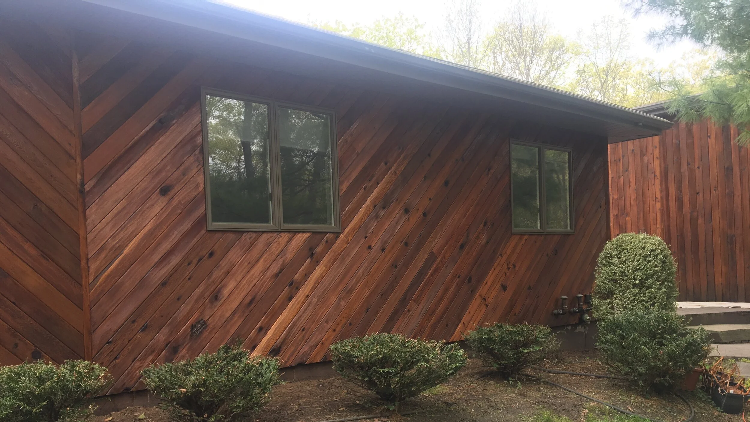 A house with diagonal wooden siding, two rectangular windows, and a small garden with bushes in front and steps at the side.