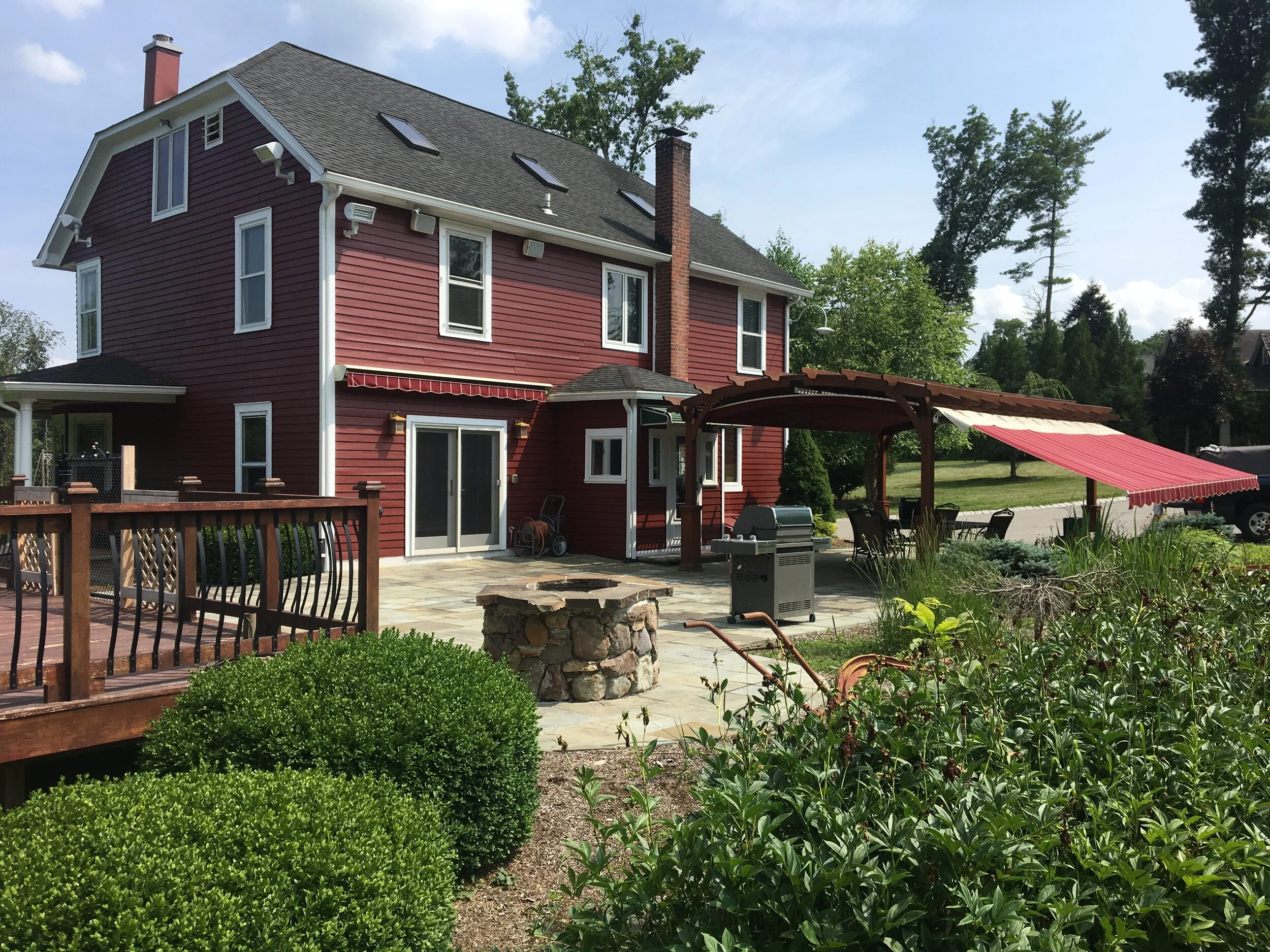 Red two-story house with white trim and a gray roof, outdoor patio area with a stone firepit, barbecue grill, patio furniture under a red and white awning, surrounded by green bushes and tall trees.