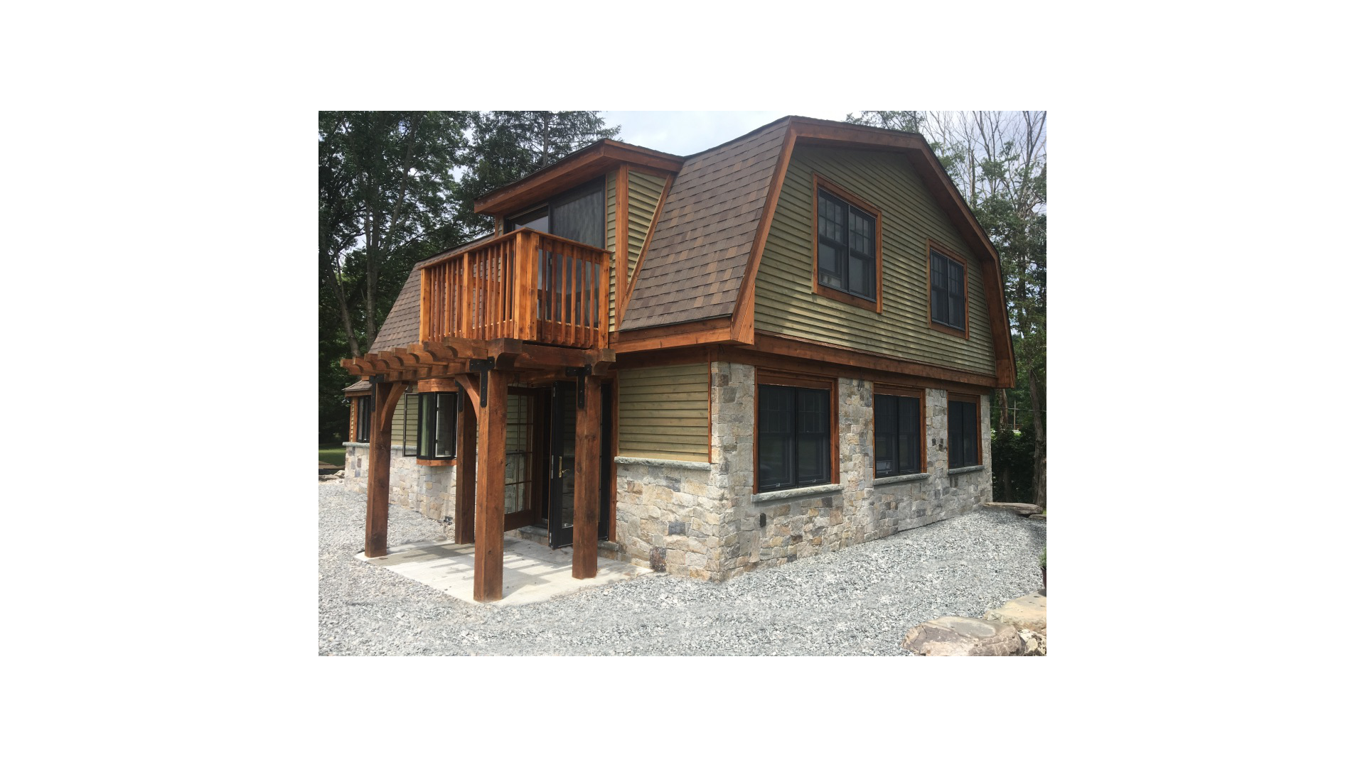 A two-story house with a stone foundation, wooden siding, and a small front porch with wooden support beams. The upper level has a balcony with a wooden railing and windows with dark frames. The roof is a combination of gable and shed styles surround