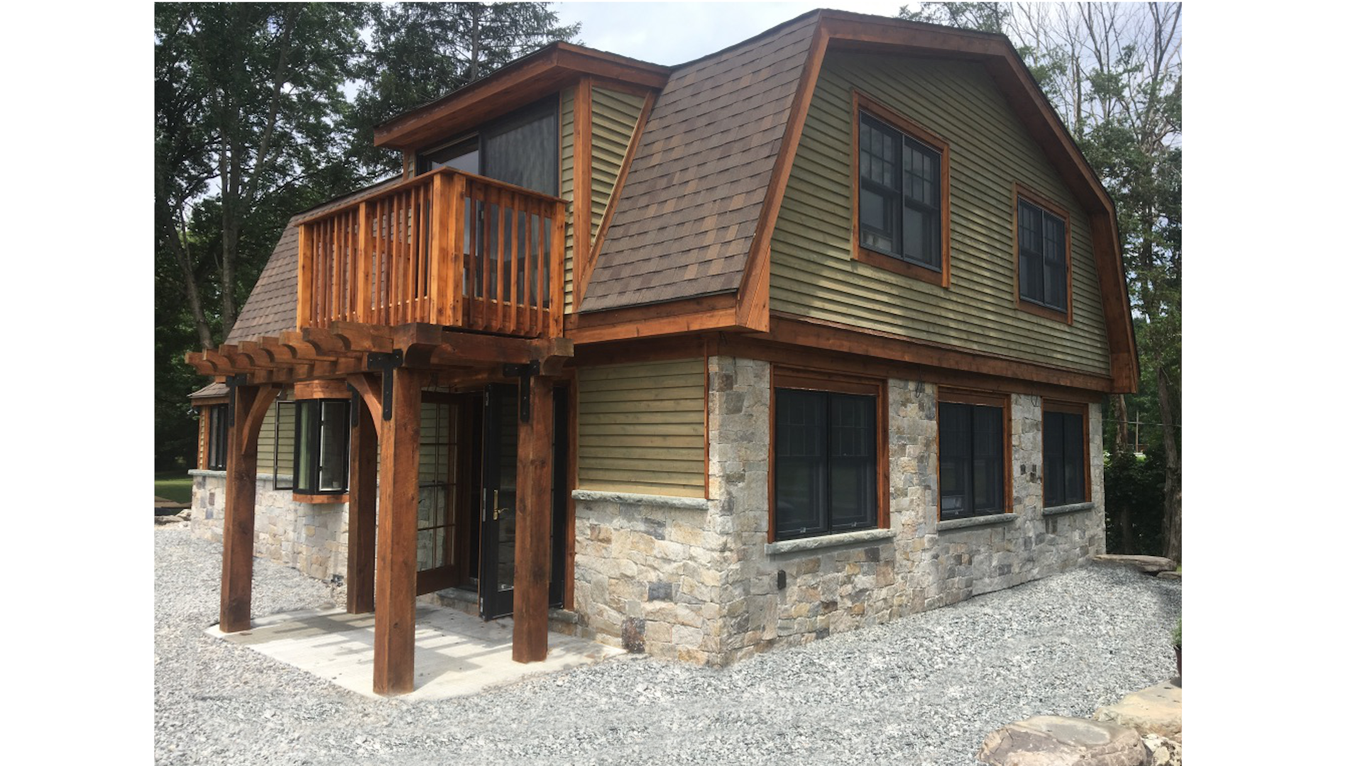 A two-story house made of stone and wood, featuring a small balcony, multiple windows with black frames, and a gravel yard surrounding it.