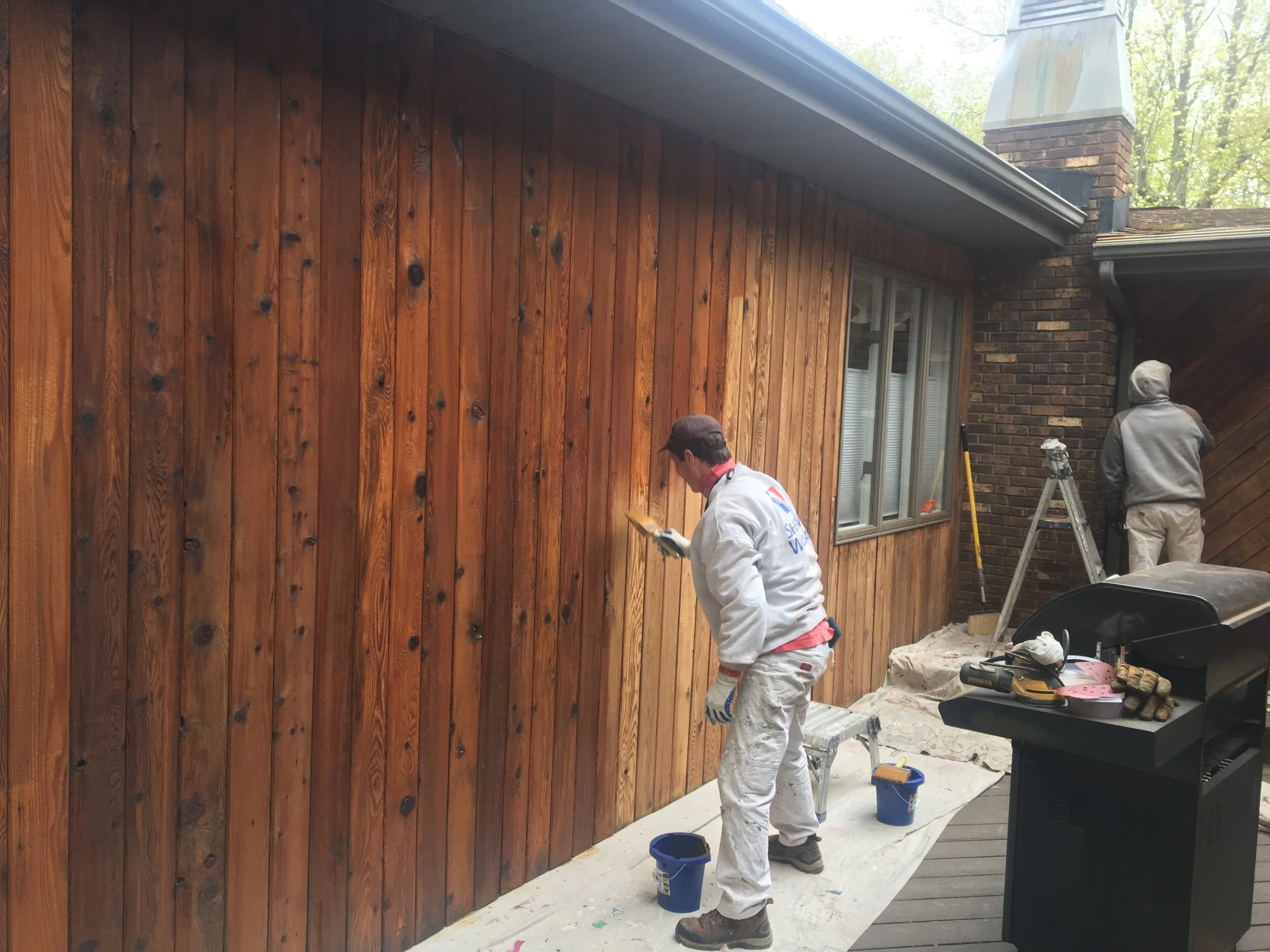 Two men painting a wooden exterior wall of a house. One man is standing and looking at a clipboard, while the other is painting near a ladder. The house has a brick chimney and a window, with trees visible in the background.