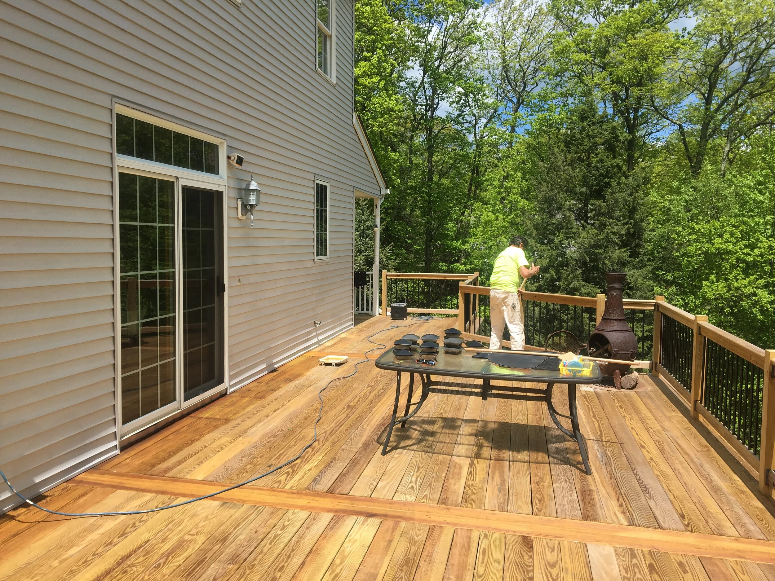 A person installing a wooden deck outside a house with sliding glass doors, surrounded by green trees in a sunny day.