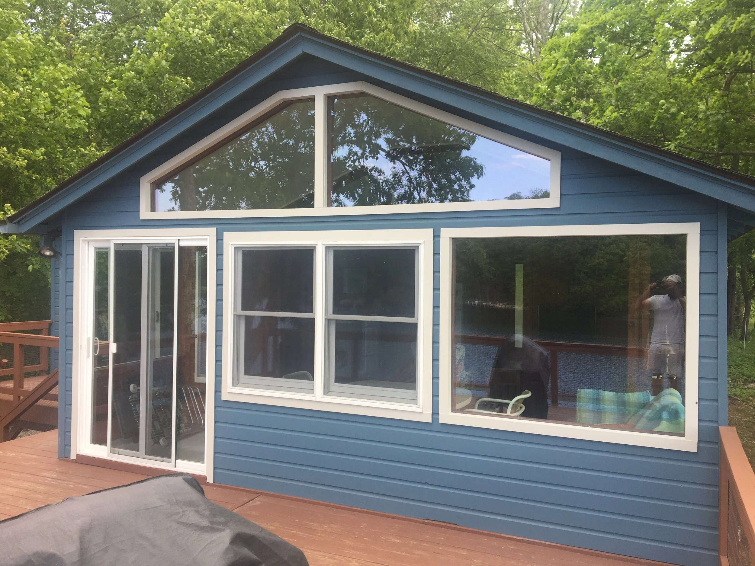 Blue house with large divided windows and a sliding glass door on wooden deck, surrounded by green trees.
