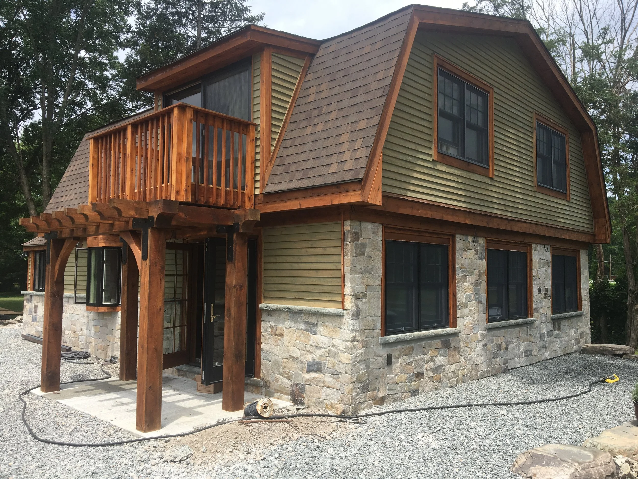 Two-story house with stone foundation, wooden siding, and a small second-story balcony with a wooden railing, surrounded by trees.